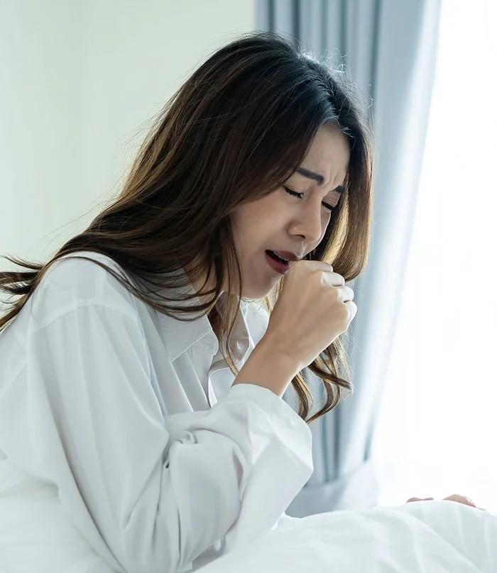 Asian woman in white sitting up in bed coughing