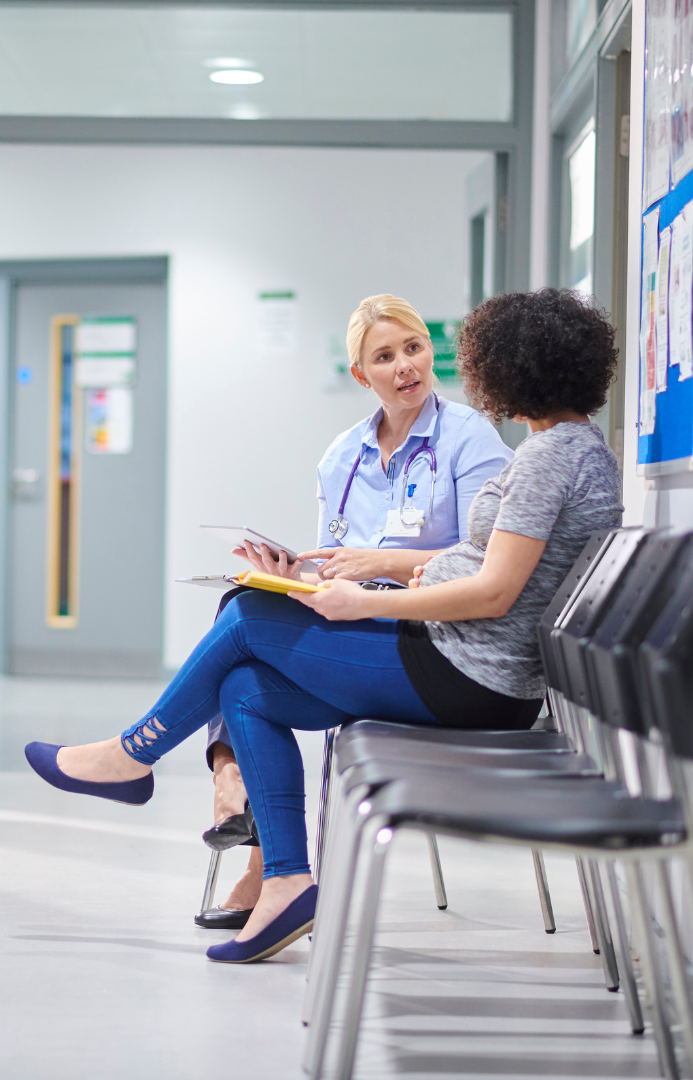 A healthcare professional, possibly a nurse, talking with a woman sitting in a hospital waiting area.