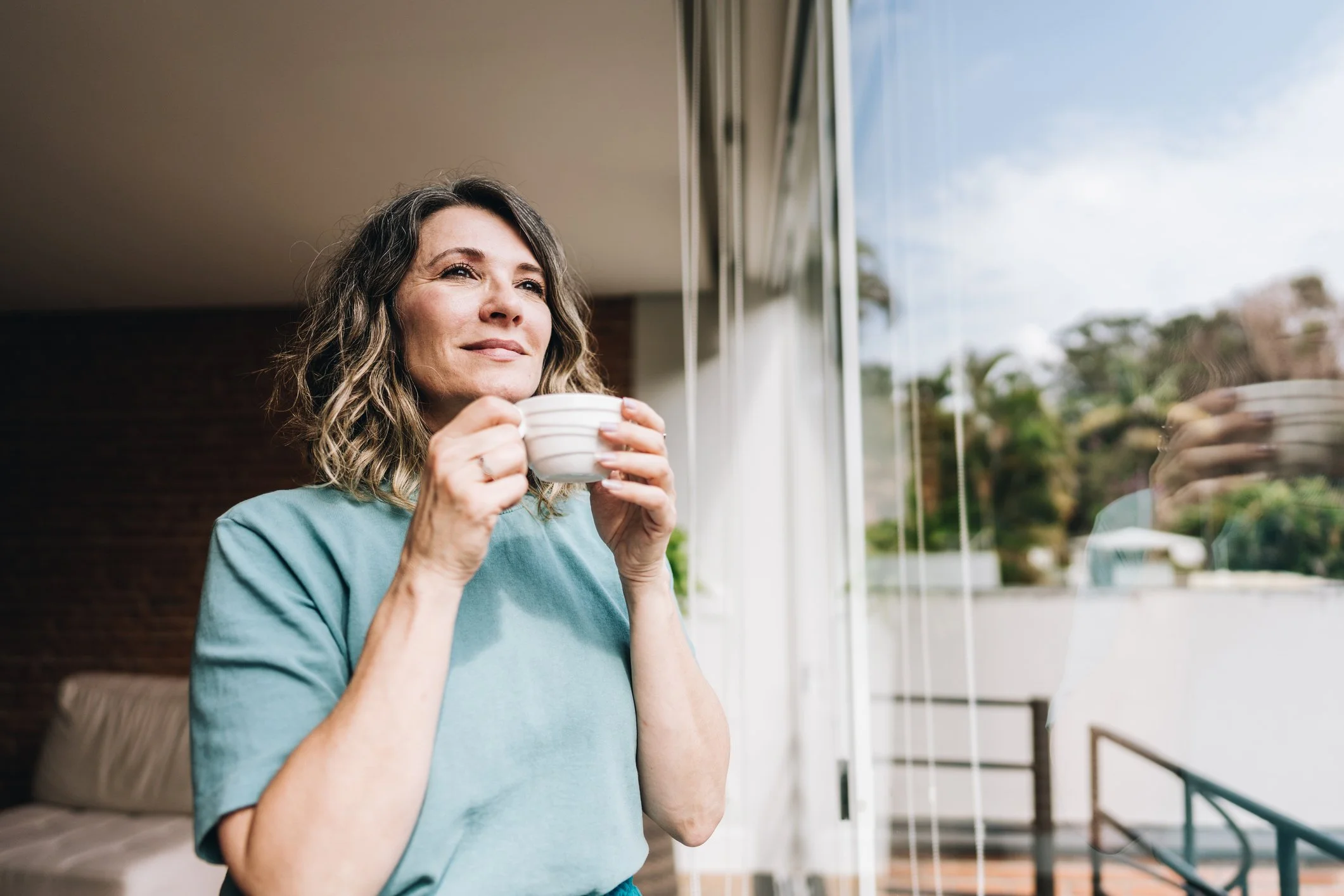 A woman holding a cup of coffee looking out the window with a happy expression