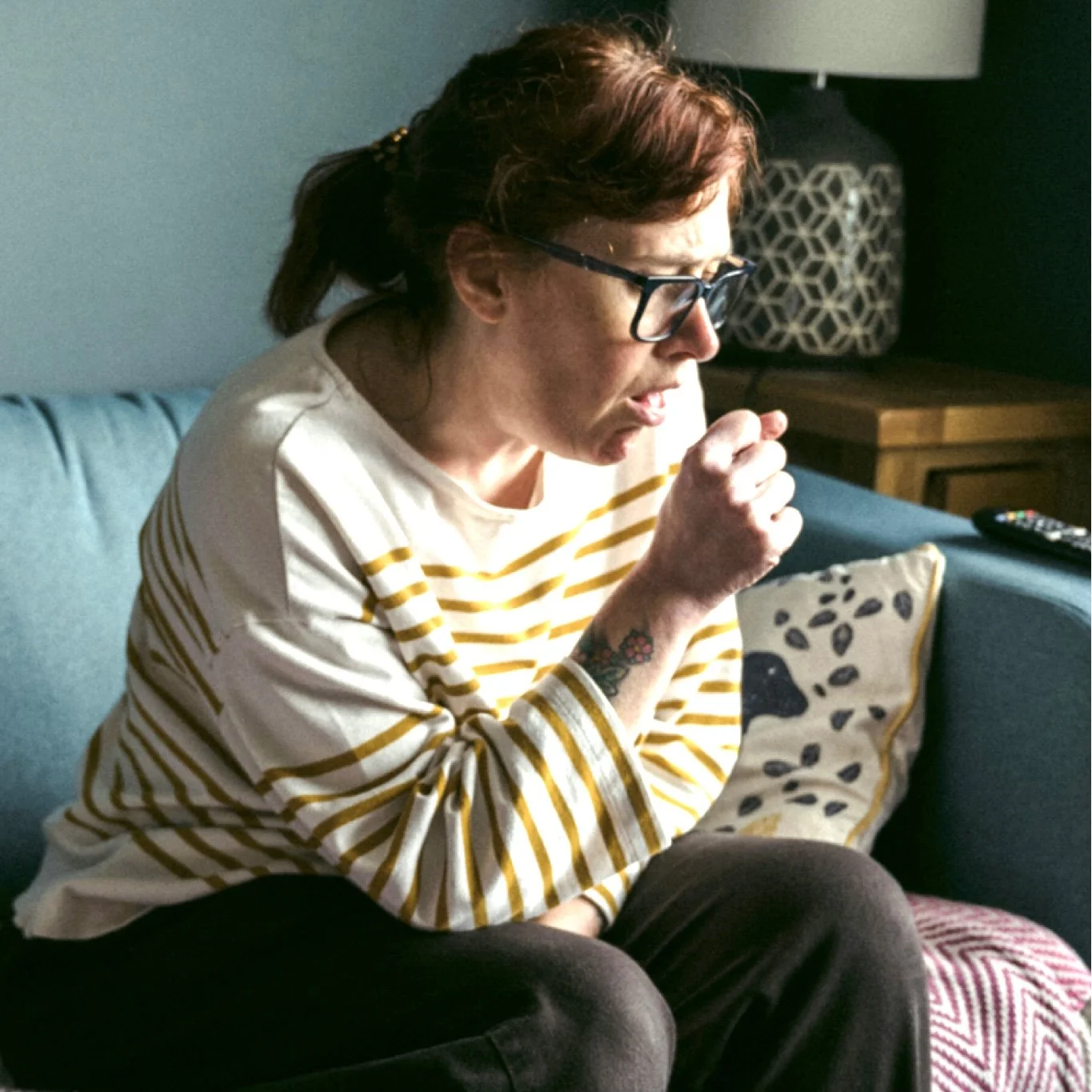 Woman sitting on sofa wearing a striped top coughing into her hand