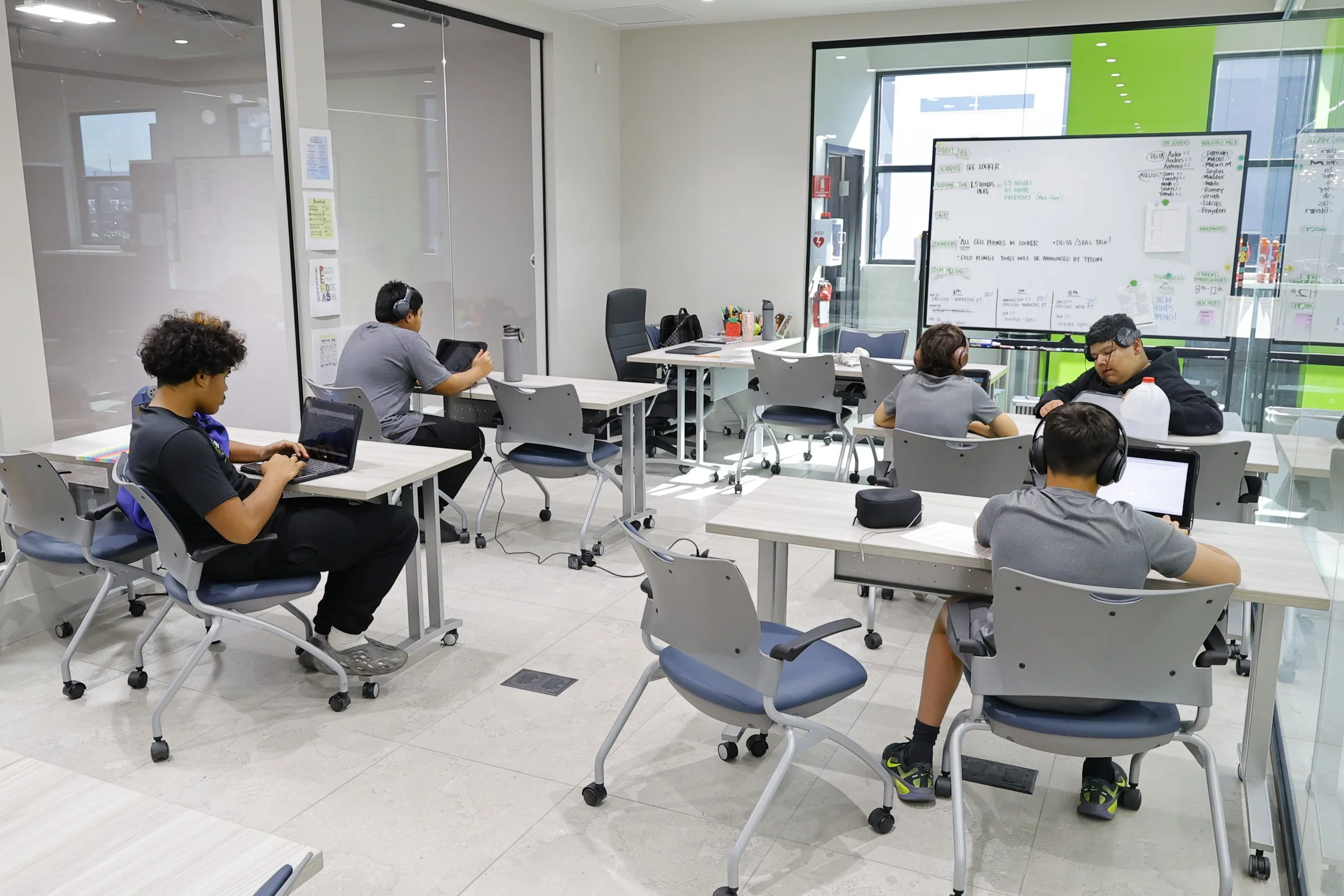 A classroom with six students seated at individual desks working on laptops, some wearing headphones, with a large whiteboard at the front filled with notes and diagrams, and windows allowing natural light to enter.