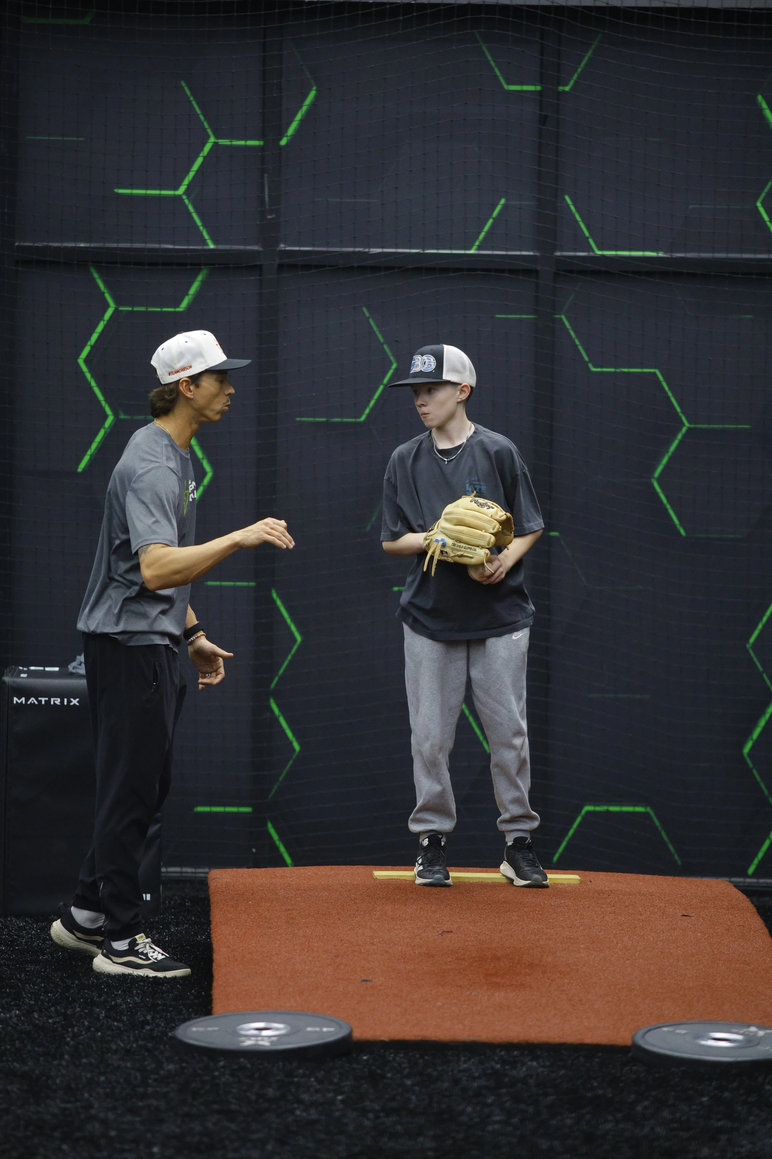 Two people standing on a practice mat inside a baseball training facility, with one person instructing and the other holding a baseball glove.