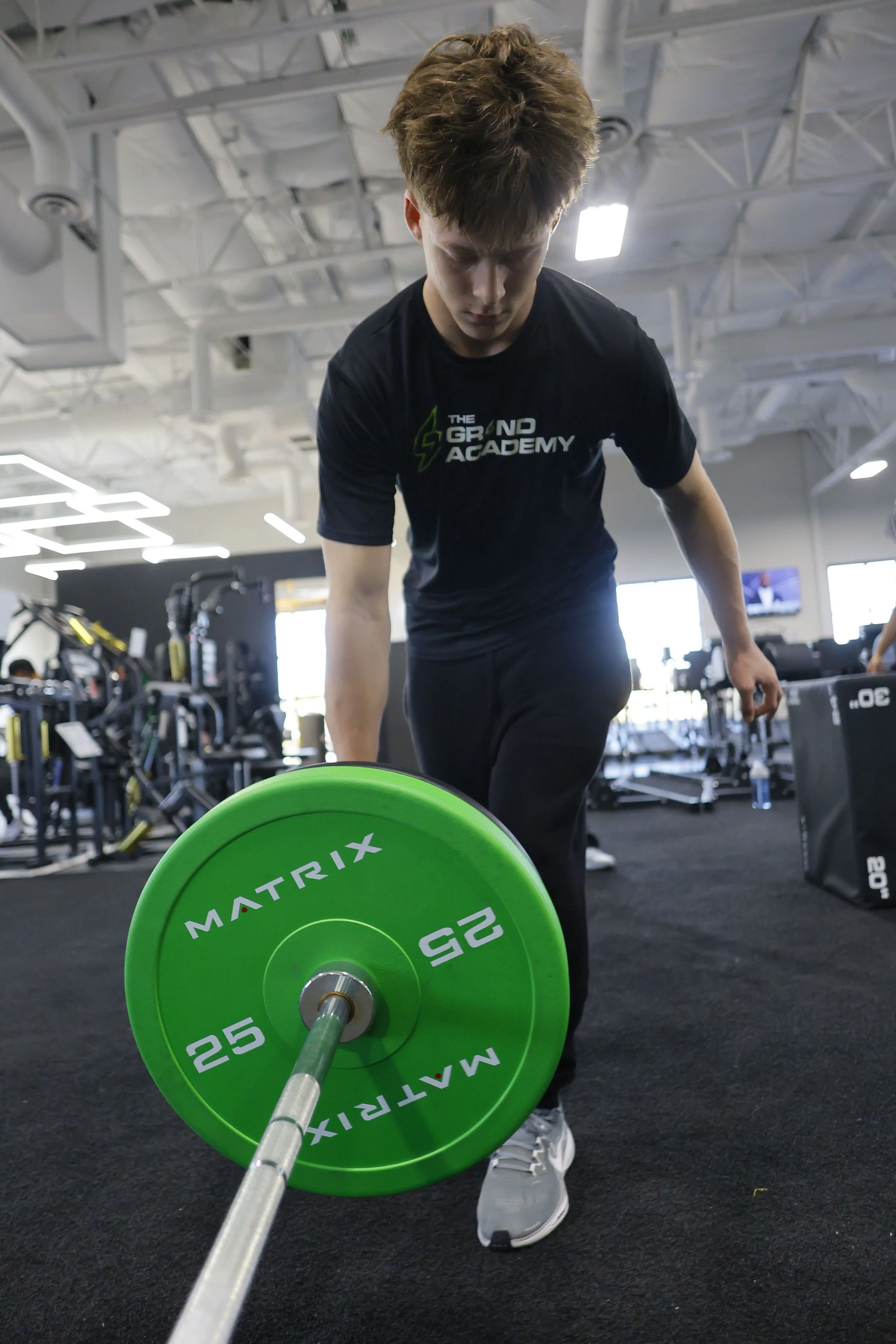 A young man wearing a black t-shirt with 'The GRIND Academy' logo is in a gym, holding a green weight plate labeled 'MATRIX 25' on a barbell, standing on a black rubber floor.