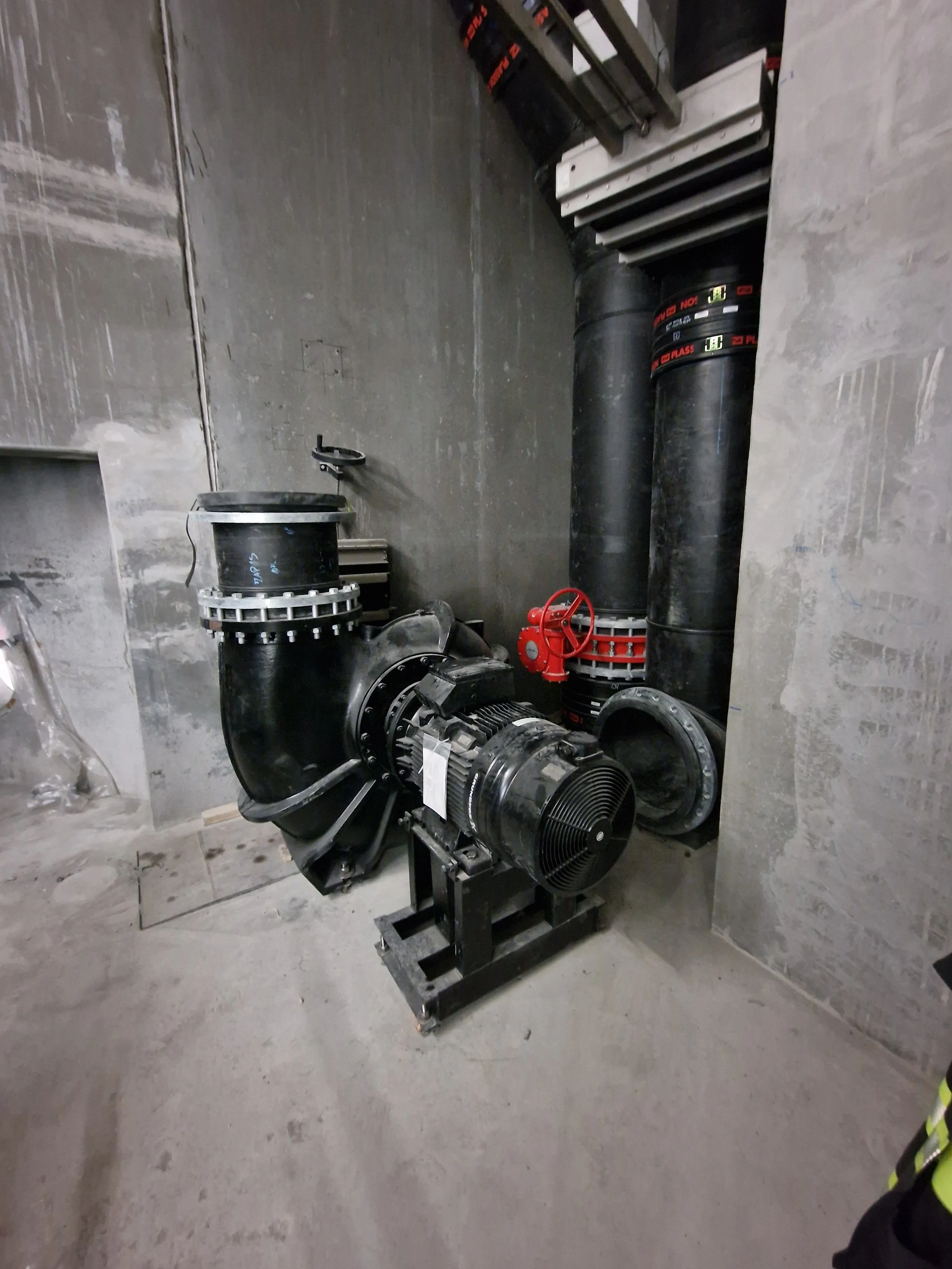En svart industriell pumpe installert i et betongrom med to store svarte rør, en rød manuell ventil, og elektriske ledninger.