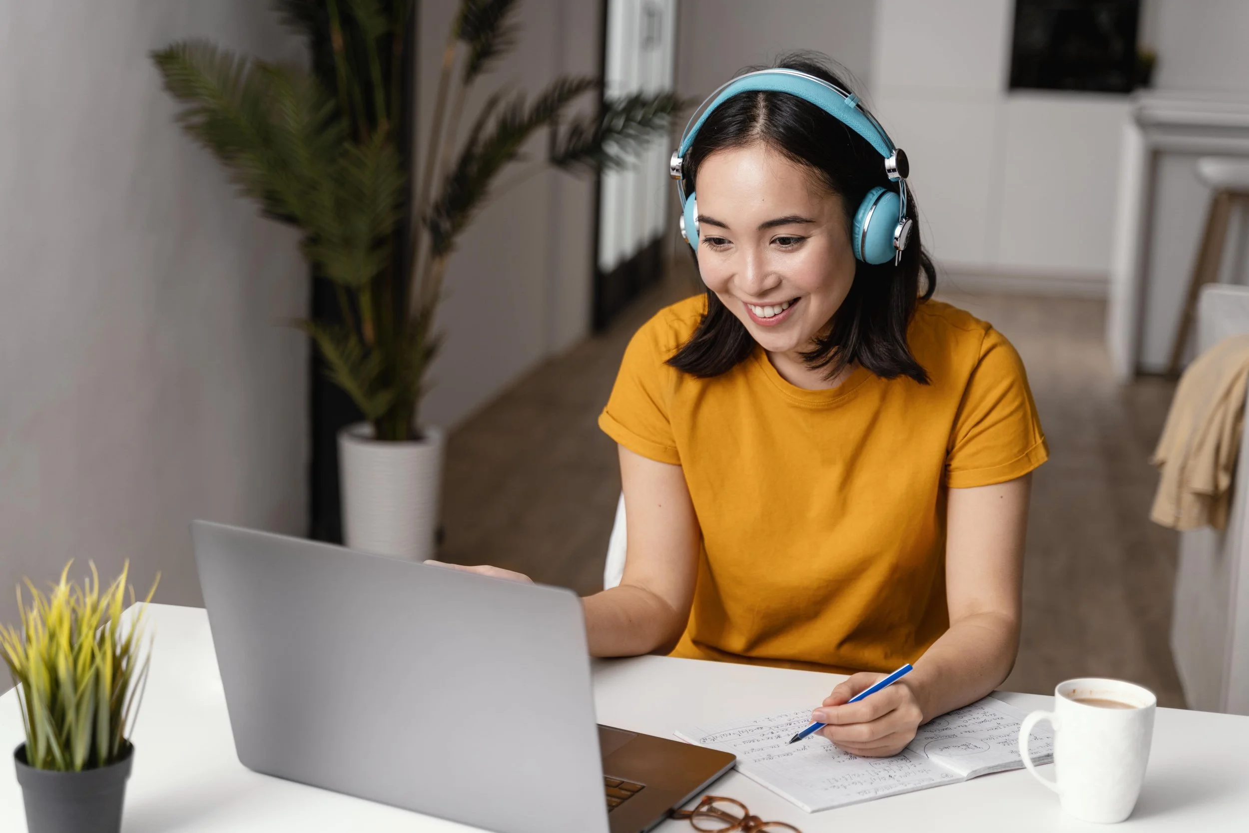 A young woman wearing a yellow t-shirt and blue headphones working at a desk, writing in a notebook with a laptop, a mug, and plants around her.