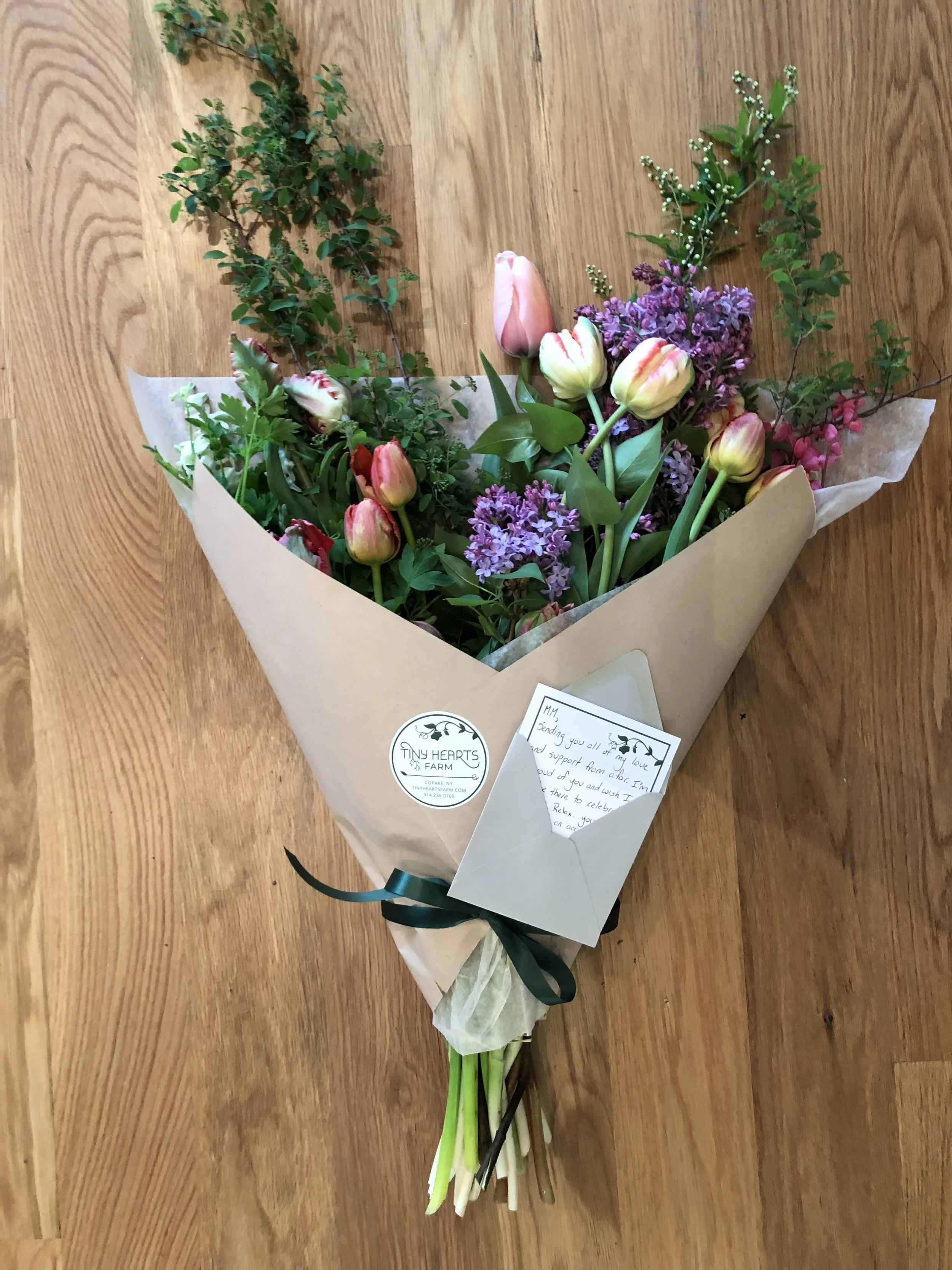 A bouquet of pink tulips, purple lilacs, and greenery wrapped in beige paper with a dark green ribbon, placed on a wooden surface.