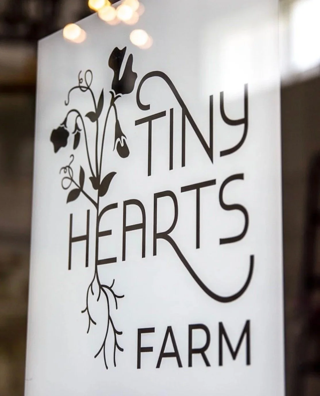 Sign reading "Tiny Hearts Farm" with a graphic of a plant with flowers and roots, black text on a white background, hanging in a rustic setting.