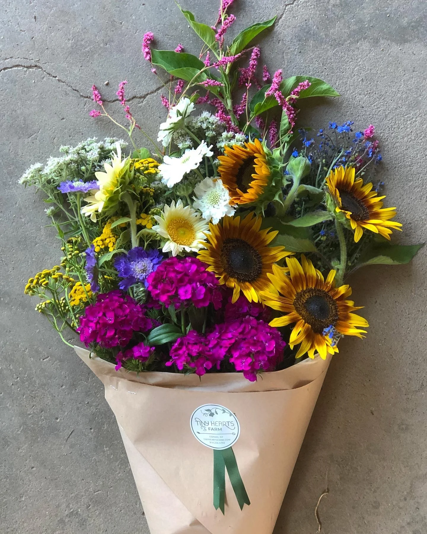 Bouquet of mixed flowers including sunflowers, daisies, and purple and pink blooms, wrapped in brown paper with a small green ribbon, label reads 'Tiny Hearts Farm'.