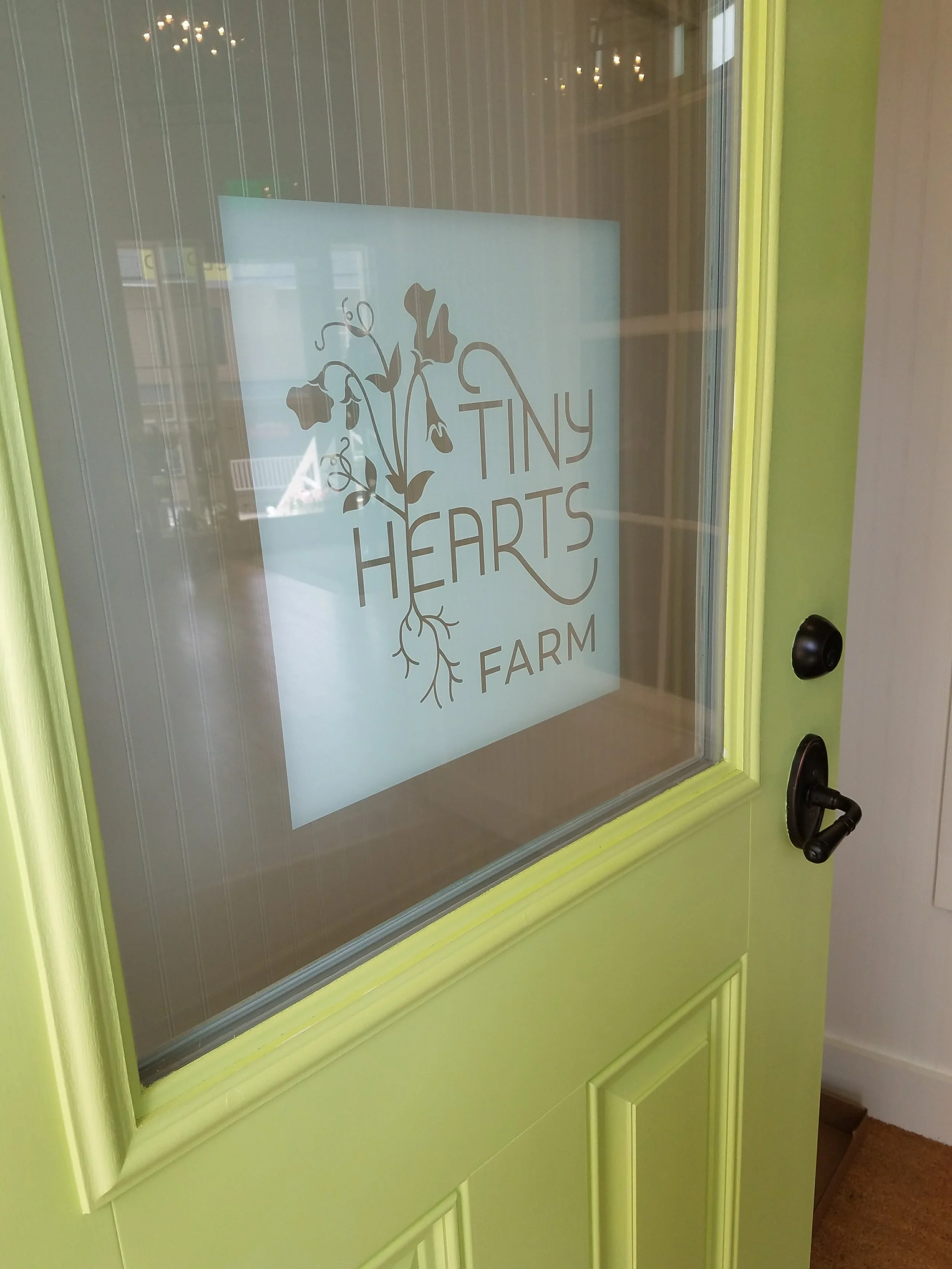 Green door with a glass window displaying a logo that reads 'Tiny Hearts Farm' with a heart-shaped plant illustration.