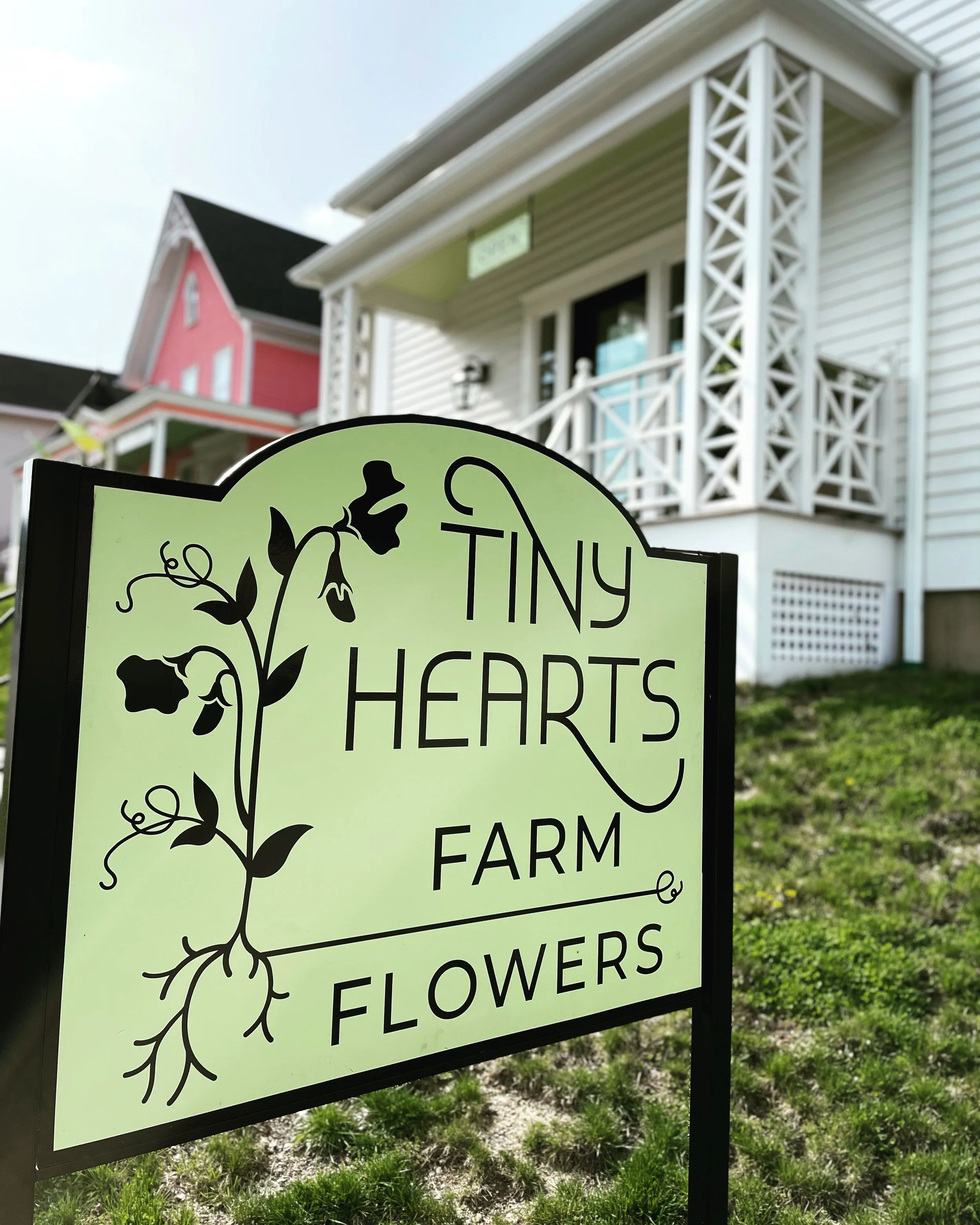 A sign for Tiny Hearts Farm Flowers with a floral graphic in front of a white house with a porch.