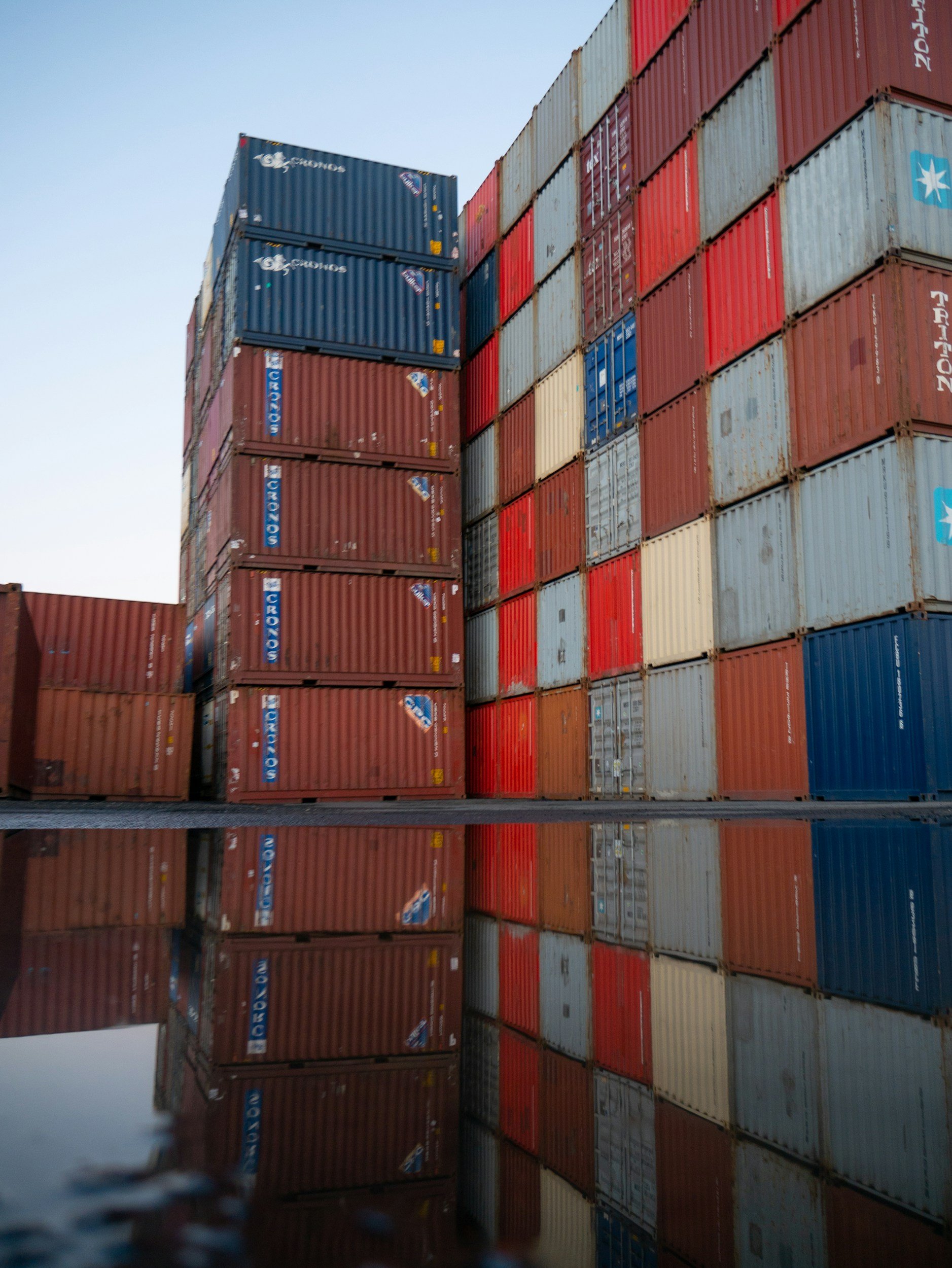Stacks of colorful shipping containers at a port, reflected in a pool of water on the ground.