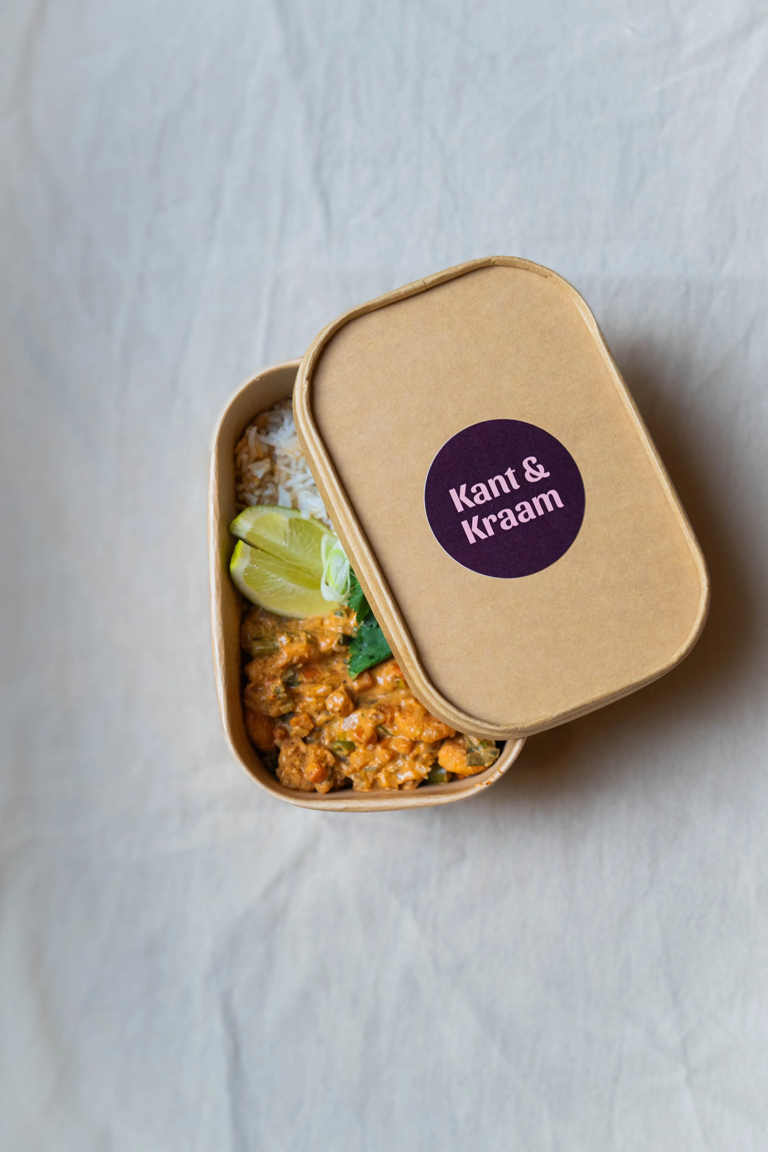 Afbeelding van een brooddoos met de woorden 'Kant & Klaar' op de deksel, gevuld met rijst, een lime, en een gekruid gerecht met groenten.