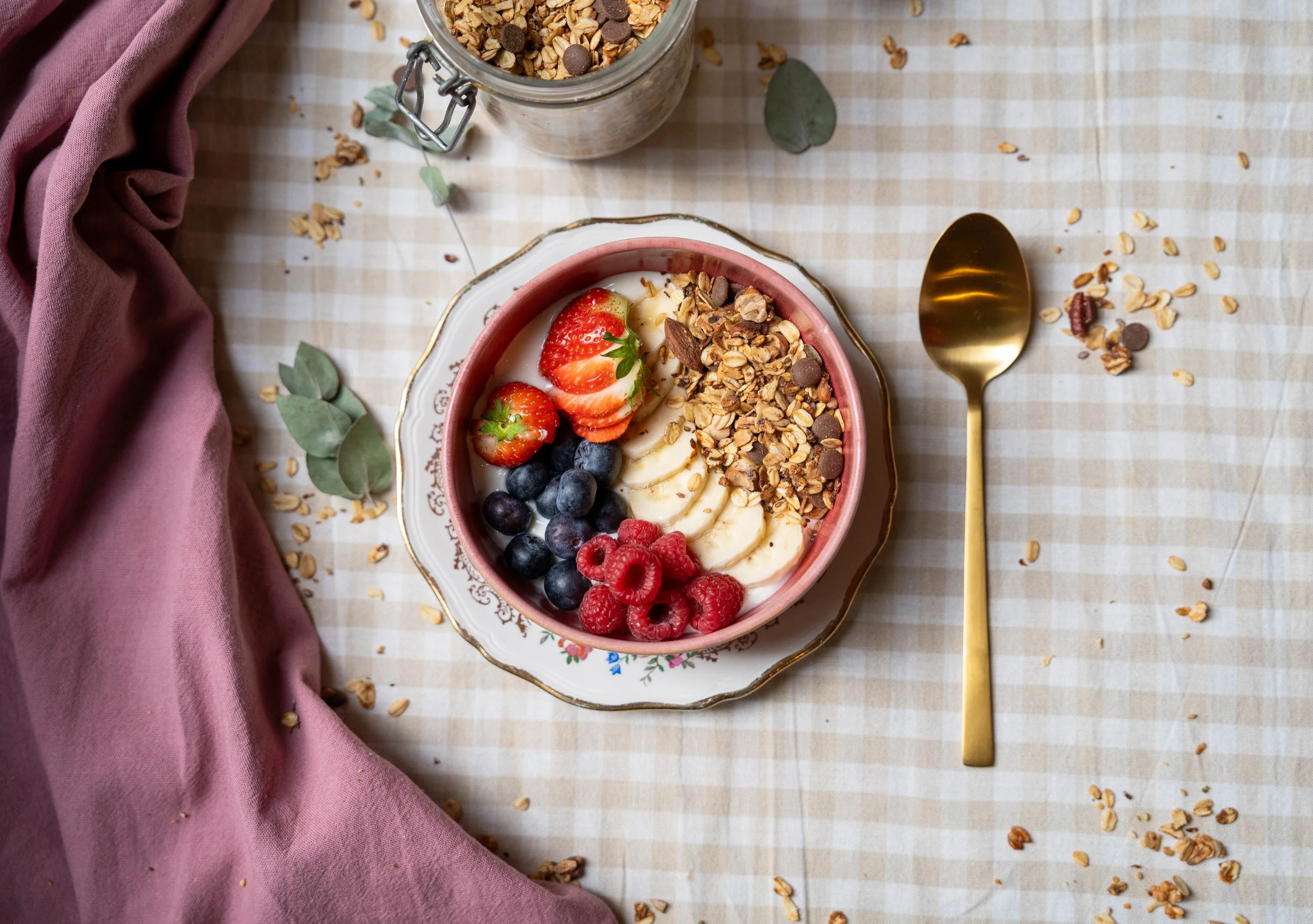 Een kom met yoghurt, besneden banaan, aardbeien, blauwe bessen, frambozen, granola en chocoladechips op een bord met een goudkleurige lepel ernaast. Er ligt ook een pot met granola en wat groene bladeren op de tafel.