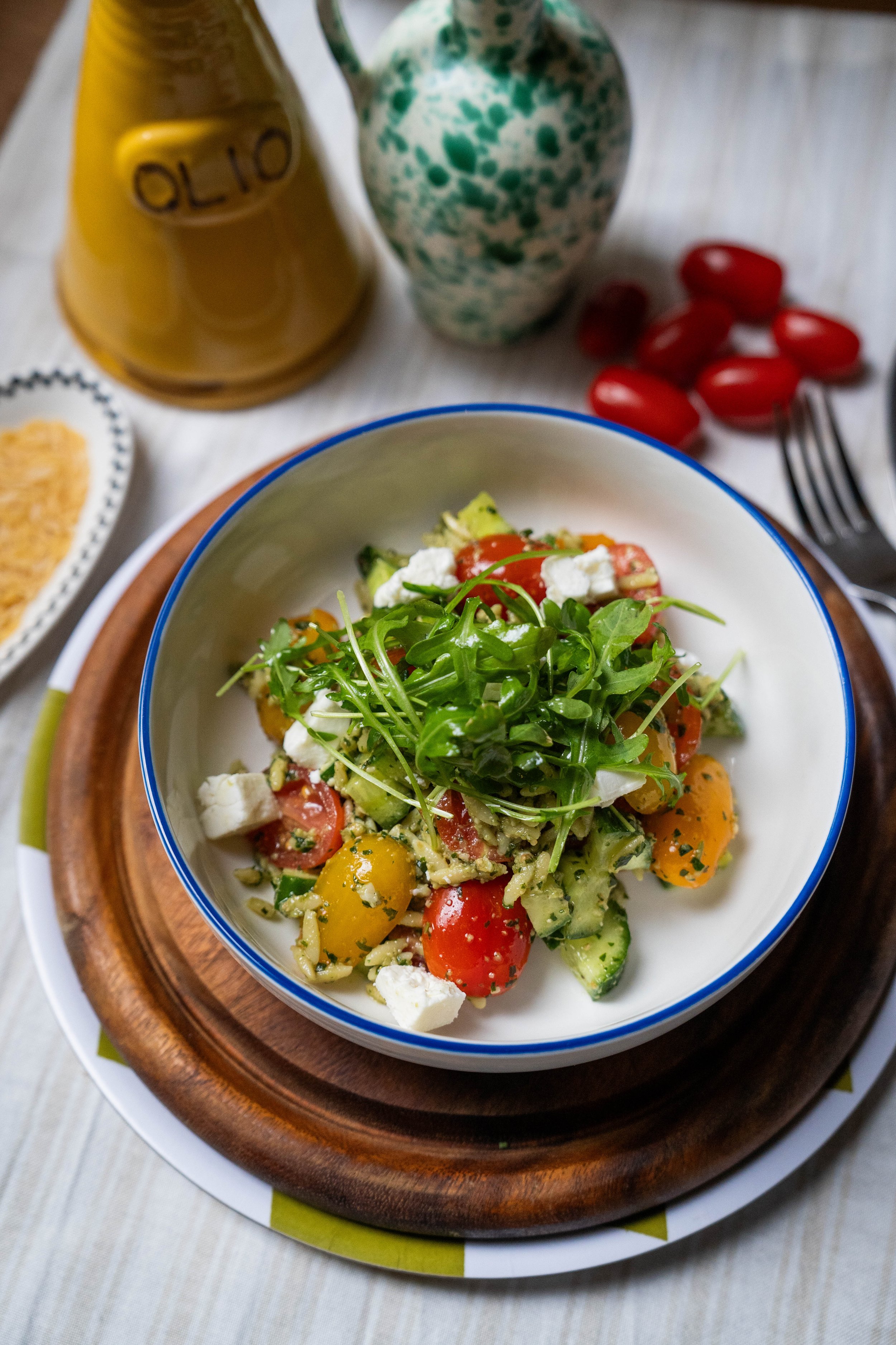 Bord met een kom salade met cherry-tomaten, avocado, rozemarijn, en witte kaas, omringd door een vork, een fles olijfolie, een vaas, en wat rode druiven.