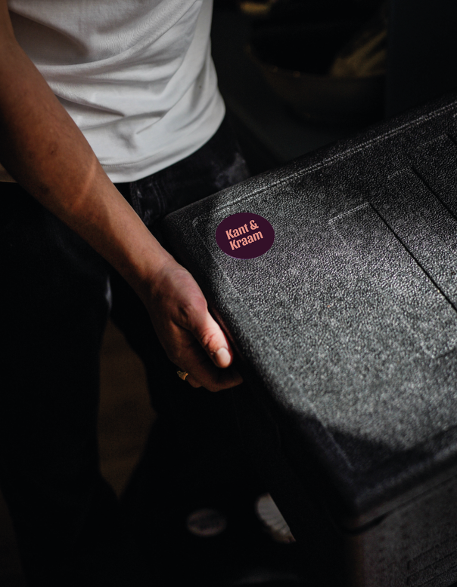 Een persoon houdt een zwart koffertje vast dat een sticker heeft met de tekst 'Kant & Kraam'.