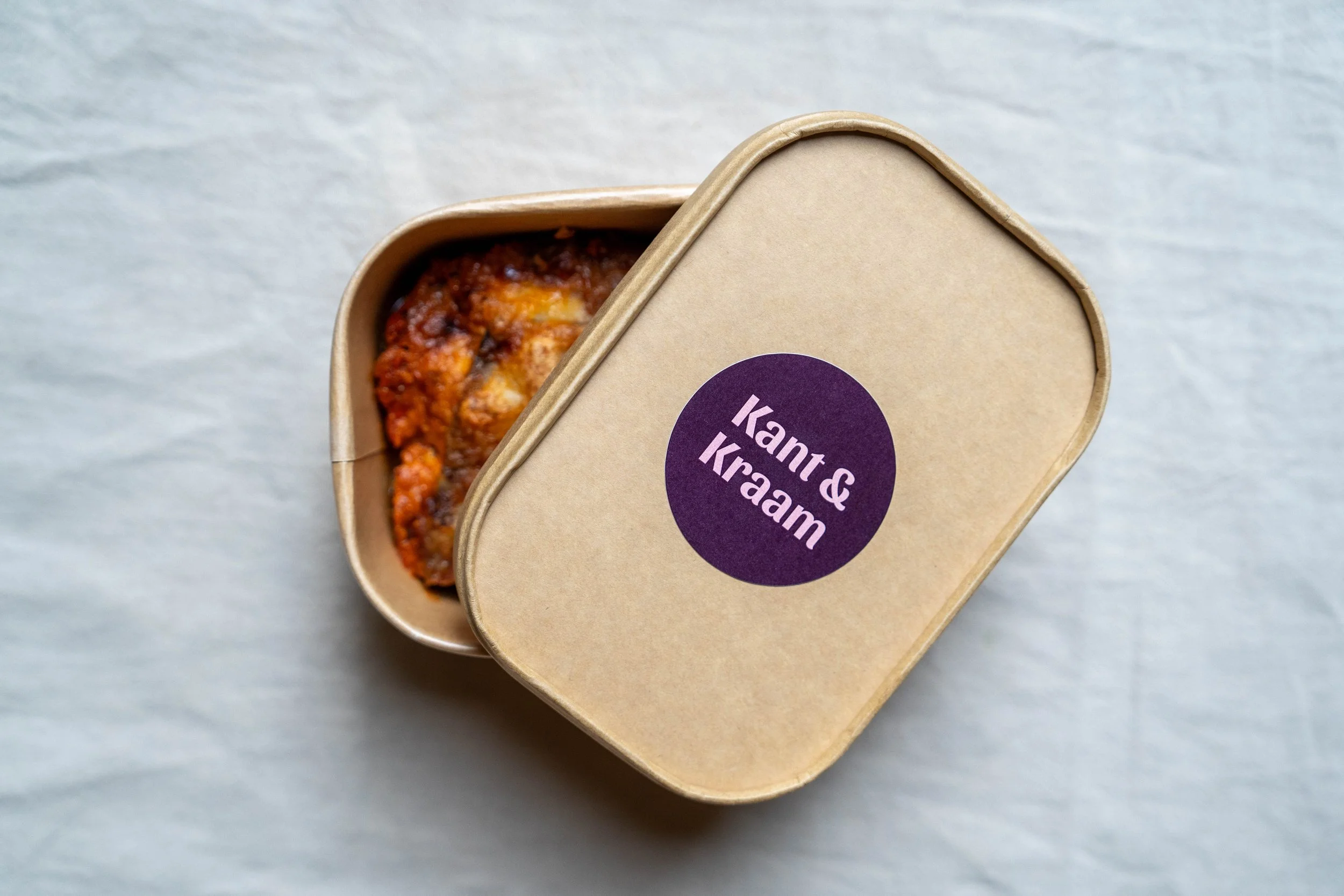 Een kartonnen doosje met de tekst "Kant & Kaam" op de deksel, waarin een ovenschotel met een saus en gesmolten kaas zichtbaar is.