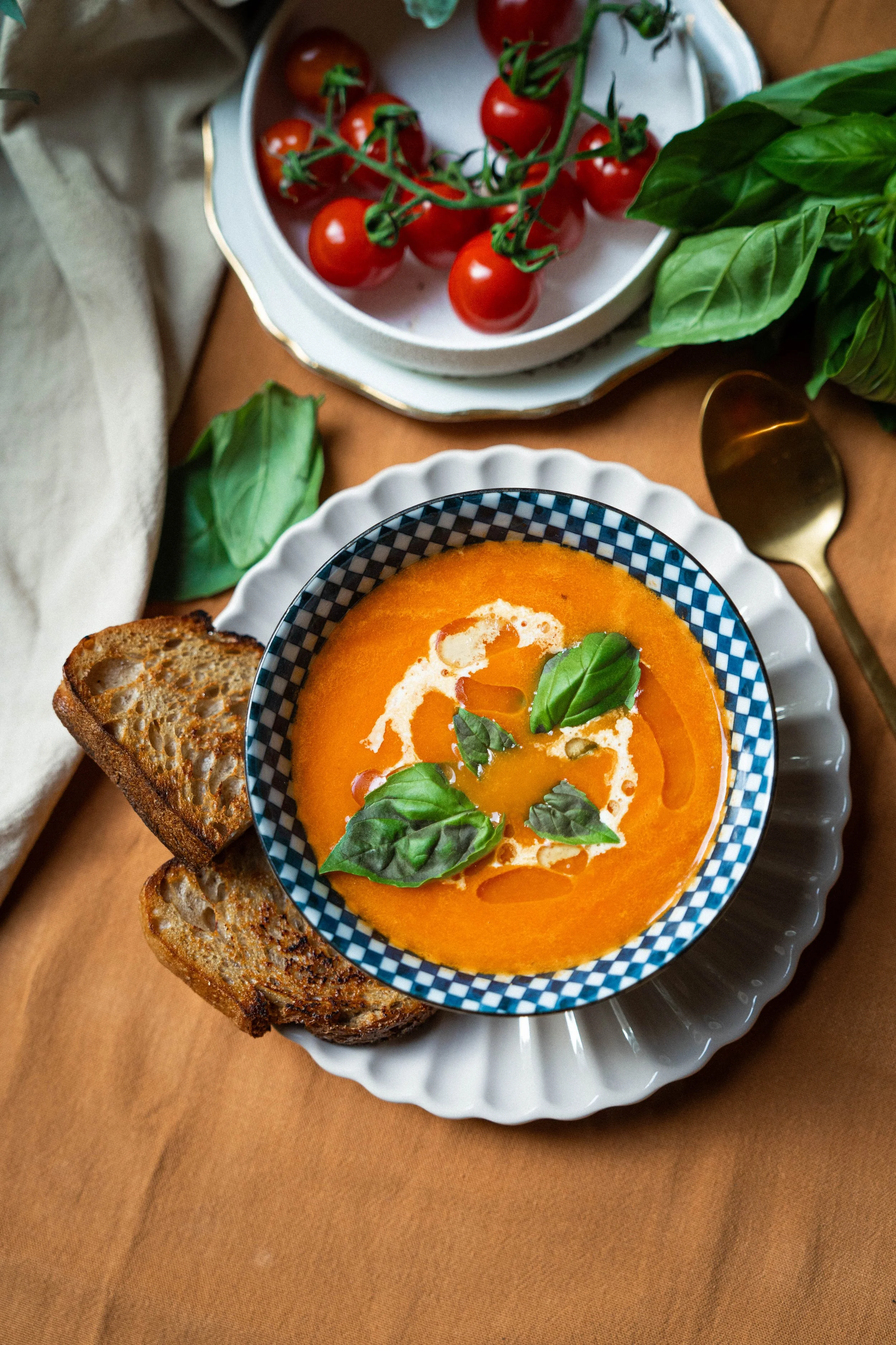 Een kom met oranje soep, versierd met basilicobladeren en een scheut olijfolie, met twee stukjes geroosterd brood ernaast.