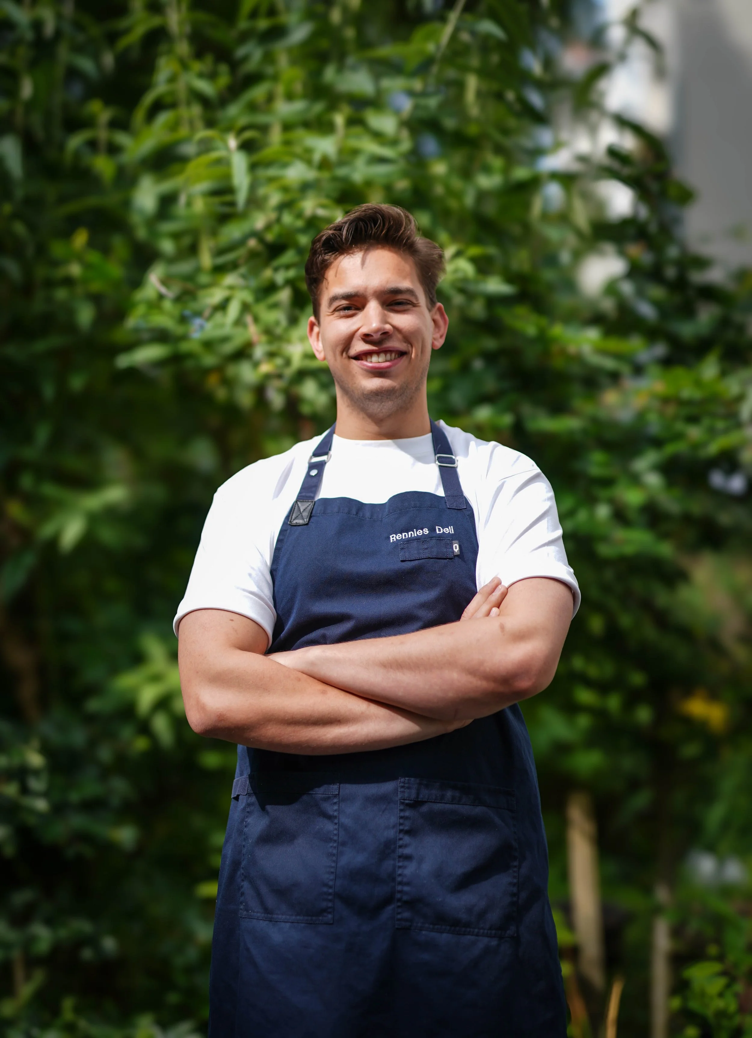 Een jonge man in een blauwe schort met de tekst 'Bennies Deli' staat met gekruiste armen voor groene planten en bomen.