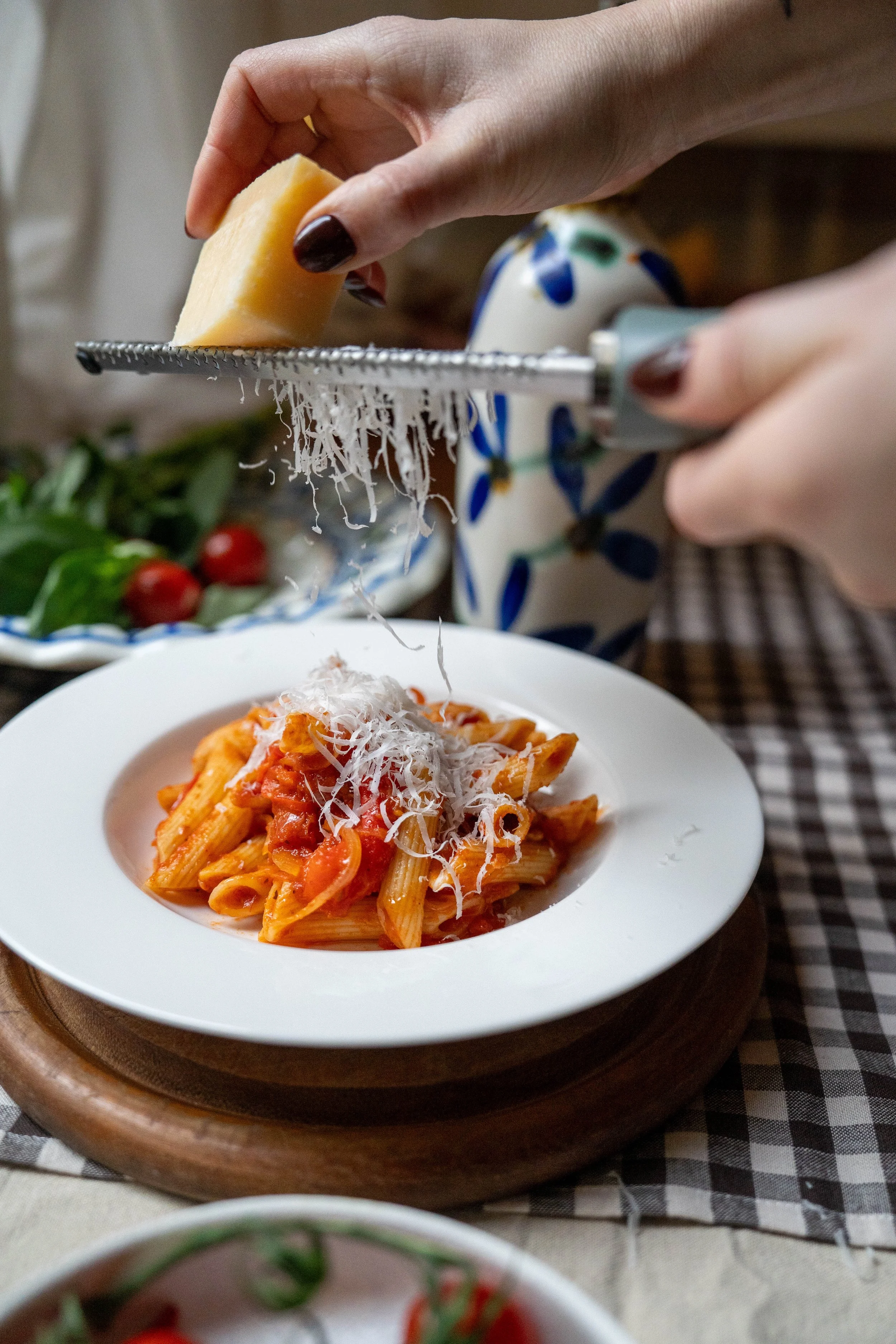 Persoon raspt Parmezaanse kaas over een bord penne pasta met tomatensaus.