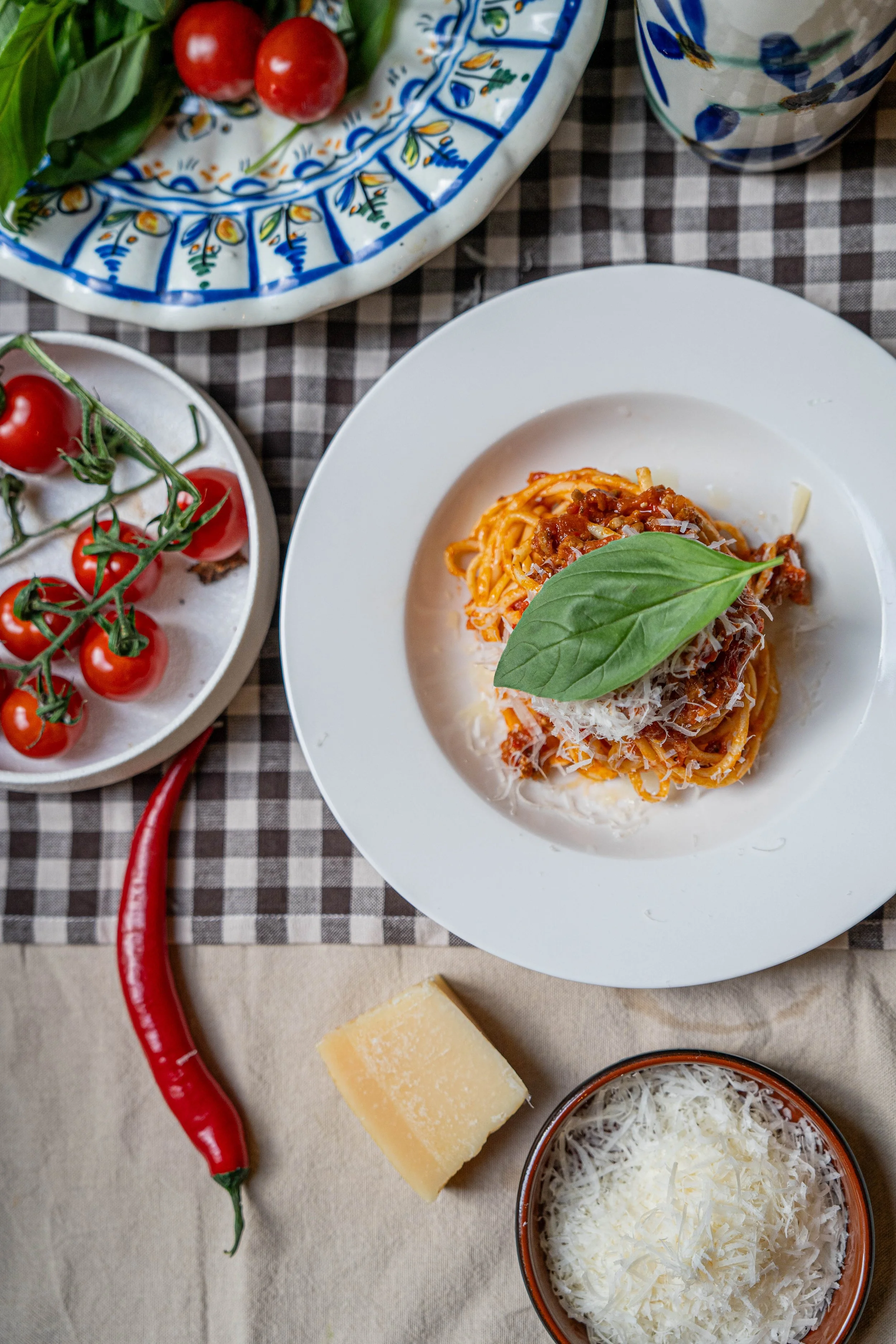 Een bord met spaghetti badden met tomatensaus en een basilicumblaadje, omringd door cherrytomaten, een stuk Parmezaanse kaas, rode chilipeper, een glas water, en een schotel met geraspte kaas.