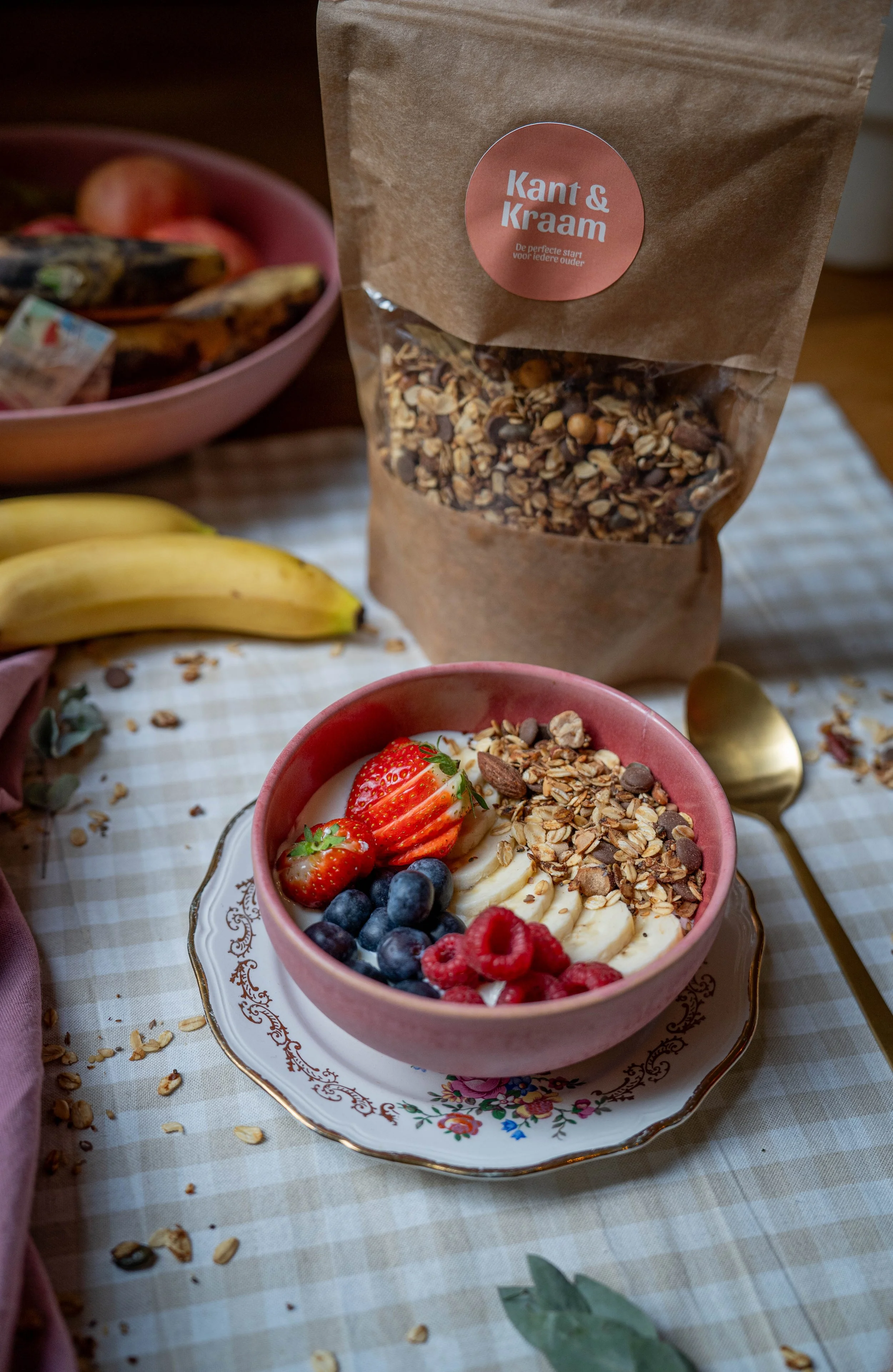 Kom met yoghurt, vers fruit, en granola, op een dienblad met een goudkleurige lepel en een papieren zak met granola. In de achtergrond liggen bananen en een roze kom met meer fruit.