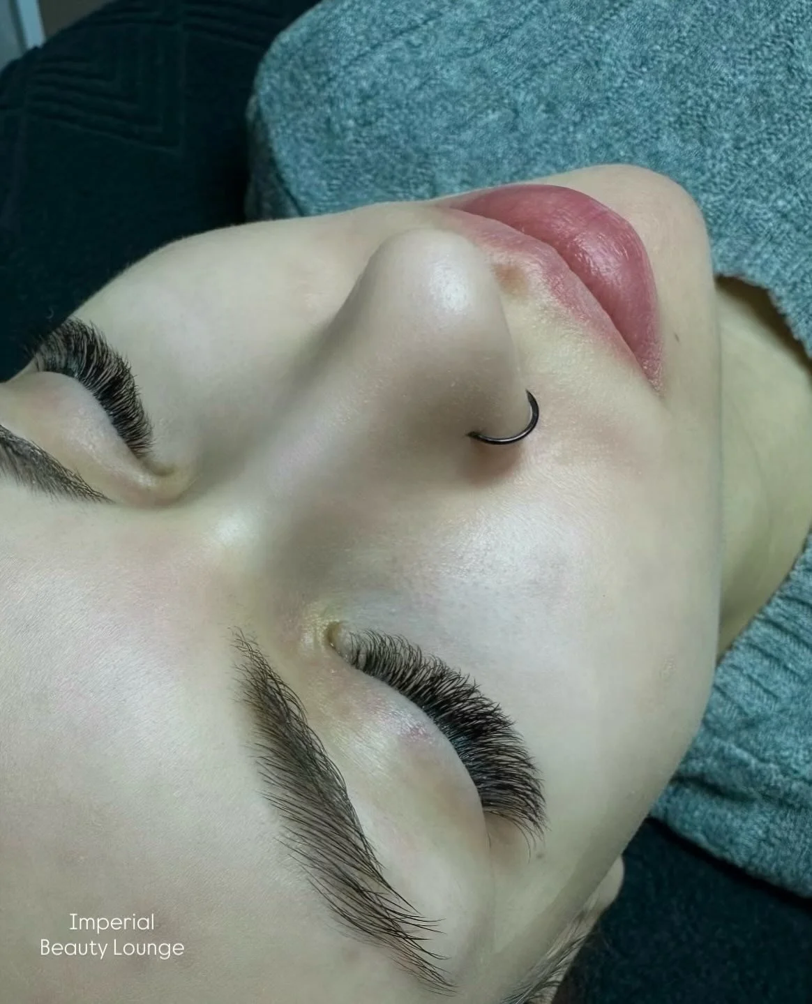 Close-up of a woman's face lying down with closed eyes, showcasing long eyelash extensions, a nose ring, and full lips. The woman appears to be at a beauty lounge, possibly after a cosmetic procedure.