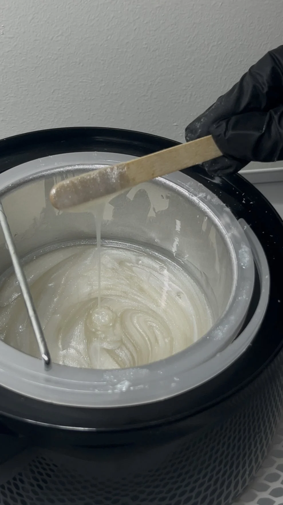 Close-up of a hand wearing a black glove stirring soap inside a large washing machine or soap maker with a wooden spatula.