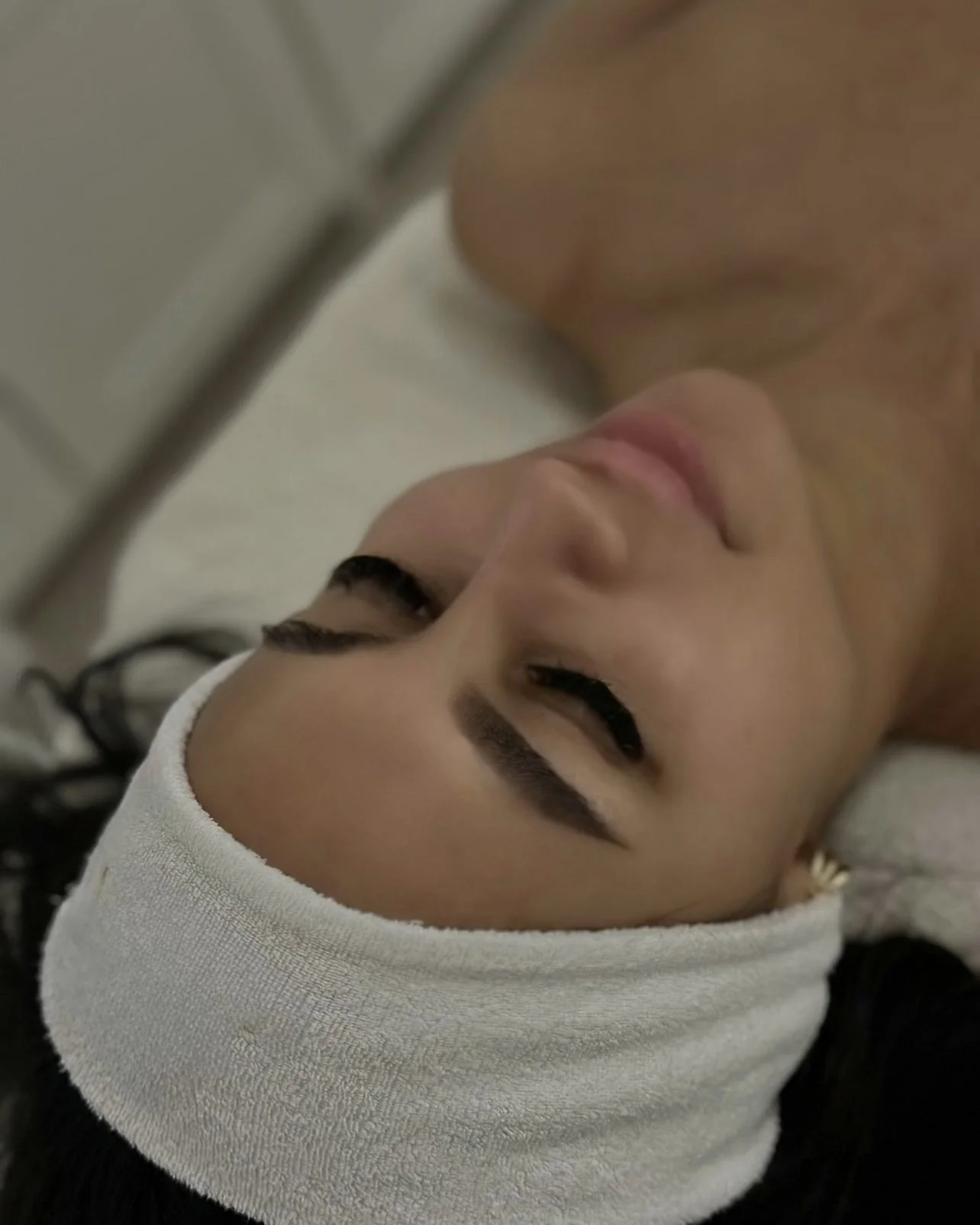 Close-up of a woman with dark hair and makeup, lying down with a white headband, eyes closed, and a hooded figure near her face.