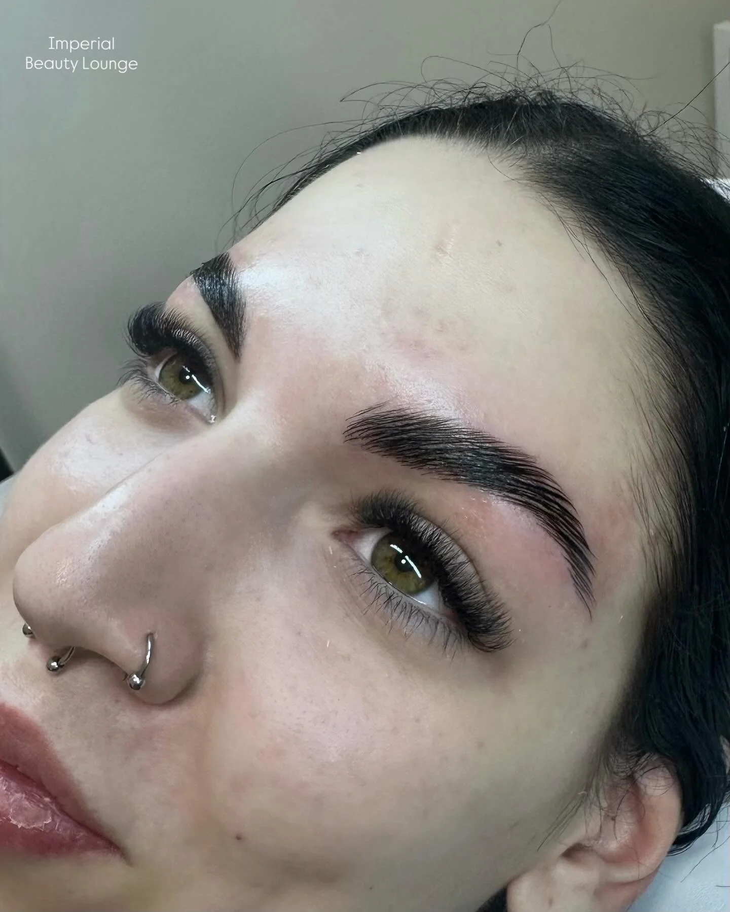 Close-up of a woman's face with freshly groomed eyebrows, a septum piercing, and subtle makeup, lying on a treatment bed at Imperial Beauty Lounge.