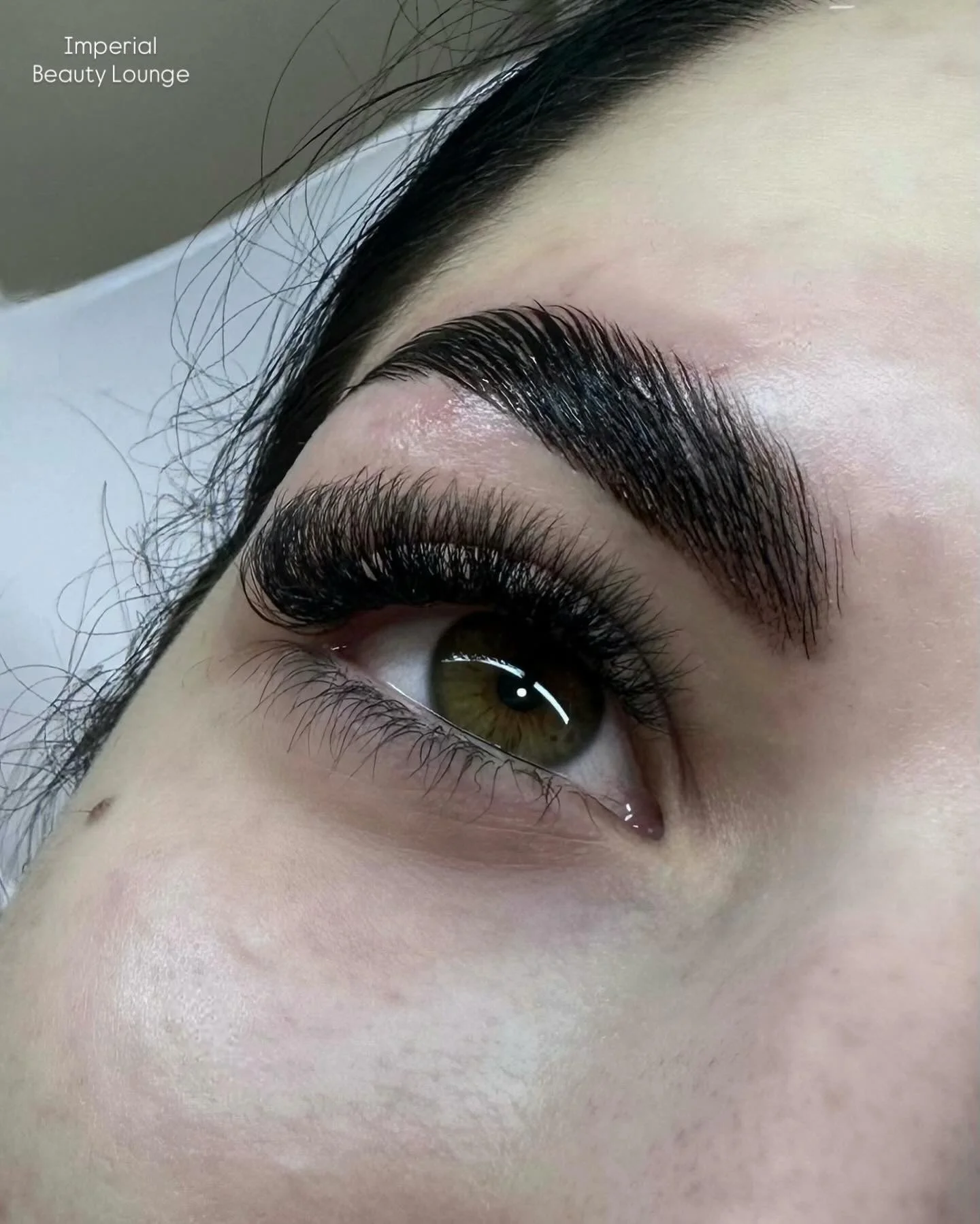 Close-up of a woman's eye with long, thick eyelashes and groomed eyebrows. The skin around the eye is smooth, with a small mole near the lower eyelid. The eye is green with a hint of hazel.