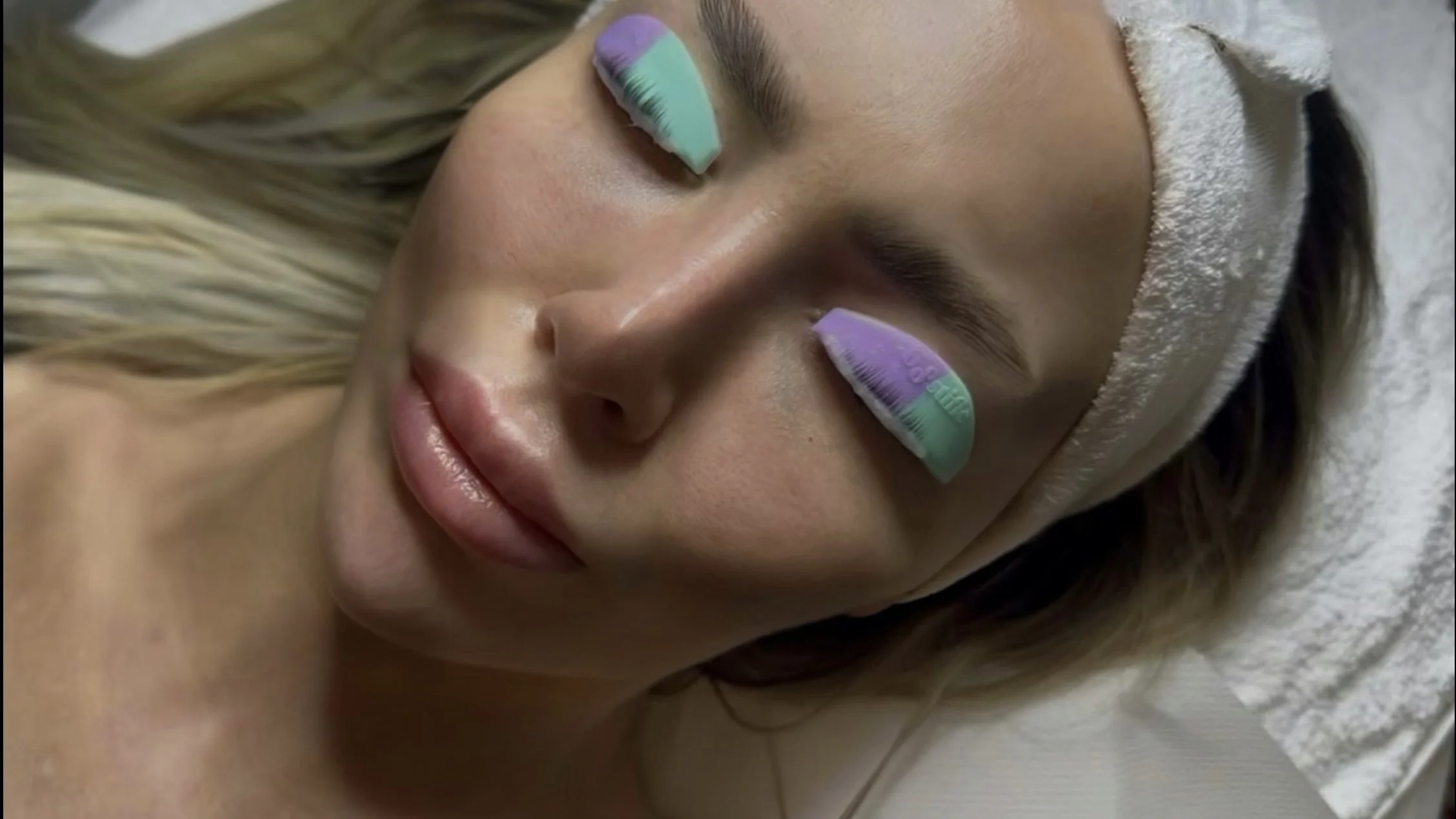 Close-up of a woman with pastel-colored eyelash extensions and a towel wrapped around her head, lying down with eyes closed.