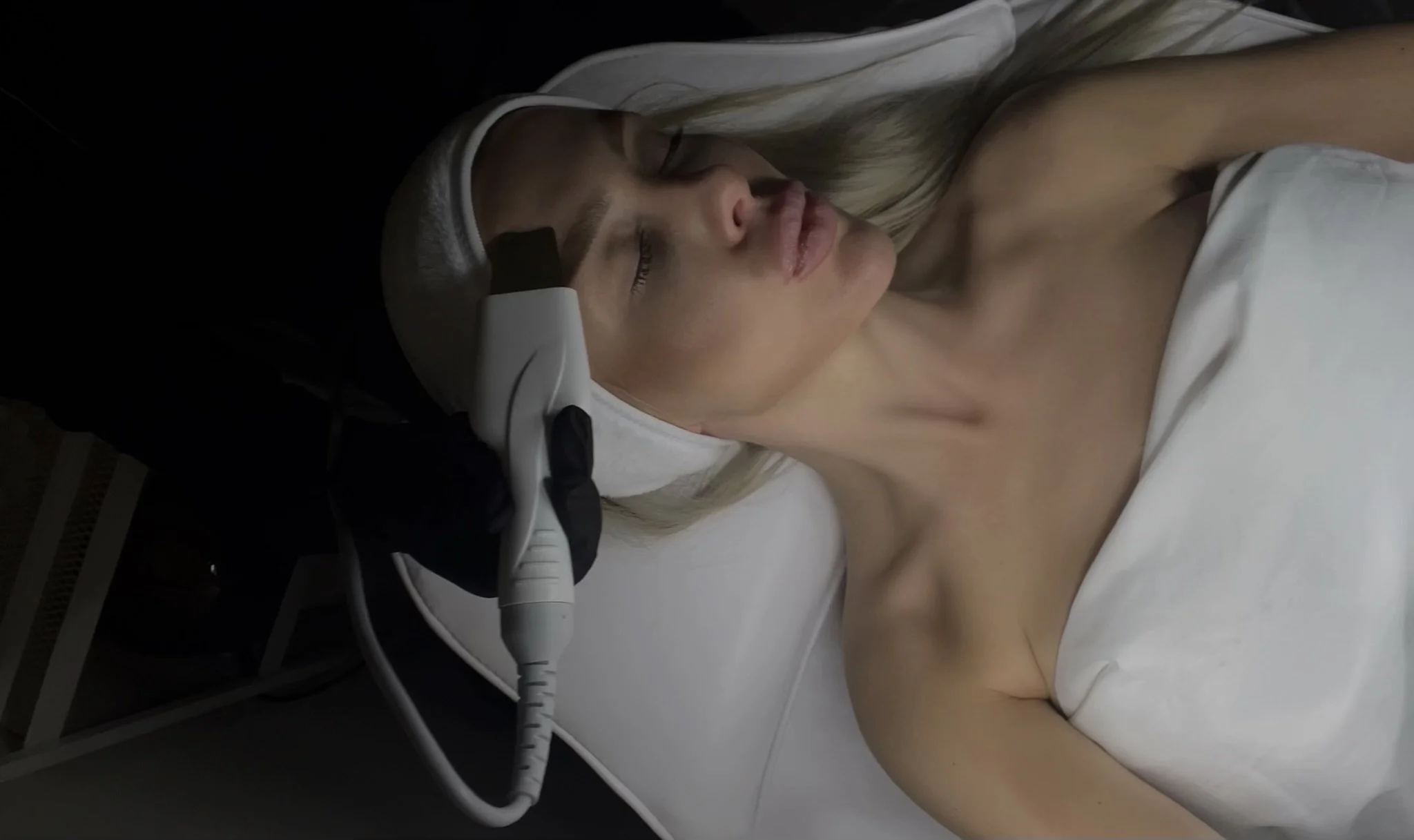 A woman with platinum blonde hair lying on a medical bed, receiving a facial treatment with a handheld device, in a clinical setting.