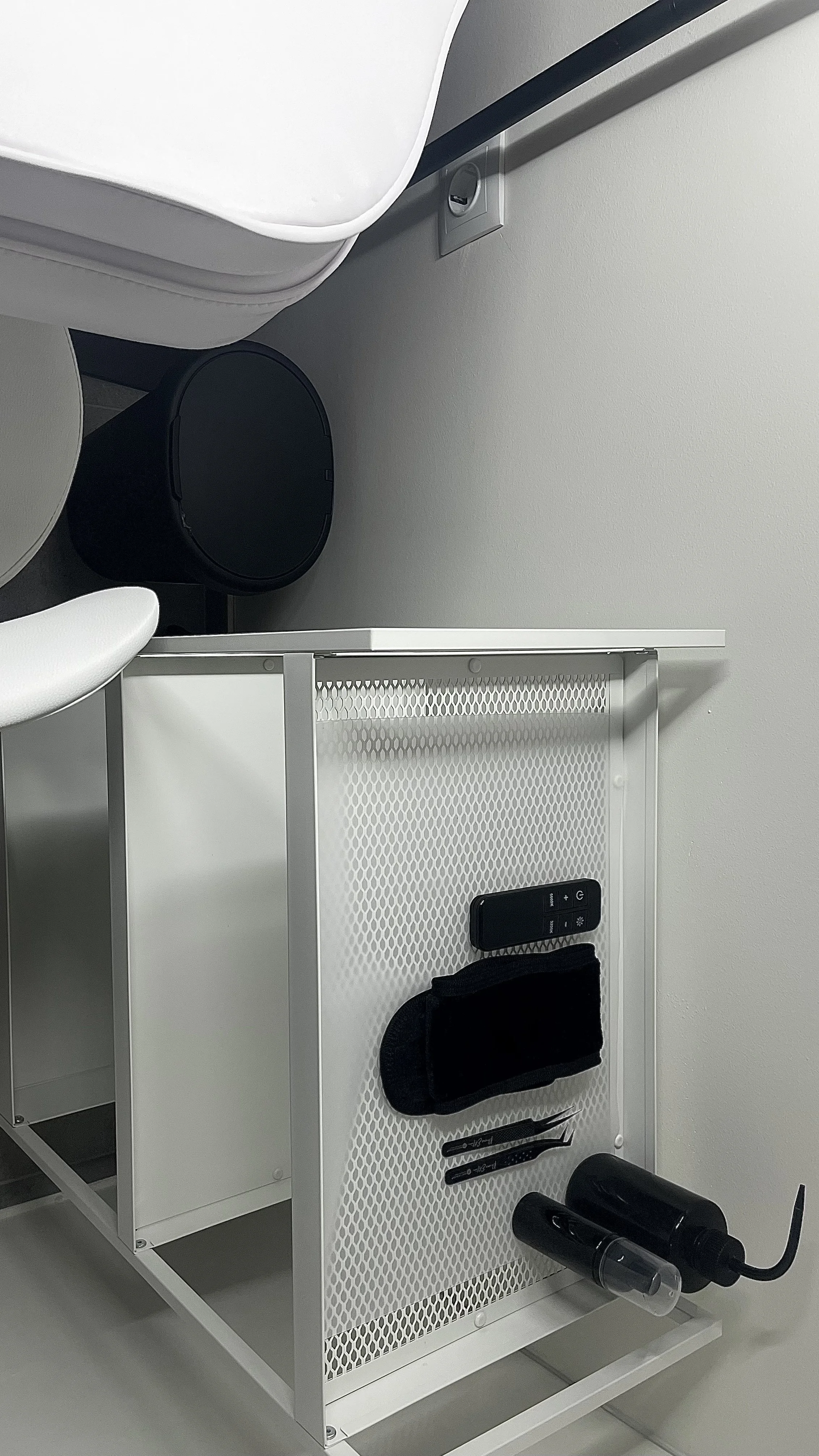 A white desk with a black speaker on top, black tweezers, and a black face mask. Next to the desk is a white mesh storage unit holding a remote control, a black vibrator, and two small black objects. The setting appears to be in a modern, minimalistic room with gray walls and a power outlet on the wall.