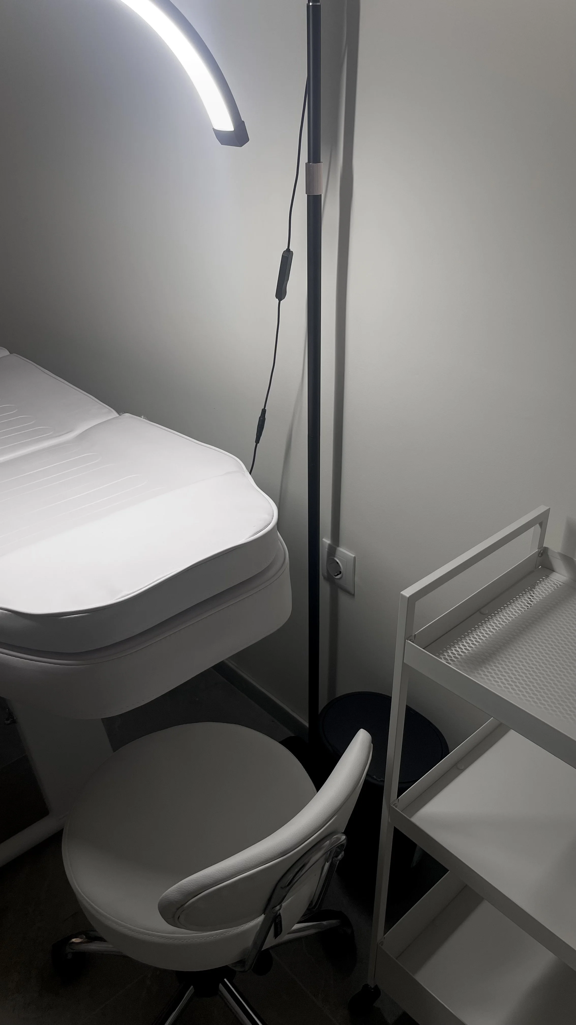 A hospital room corner with a bed, a white chair, a tall medical lamp, a three-tiered white cart, and a black waste bin, with a wall outlet and cord visible.