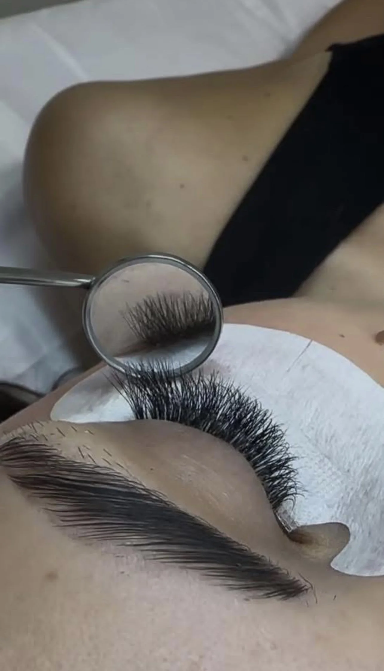 A person receiving eyelash extension application, lying on a white surface, with black eyeshadow and bold eyebrows, and a magnifying glass focusing on the new eyelash extensions.