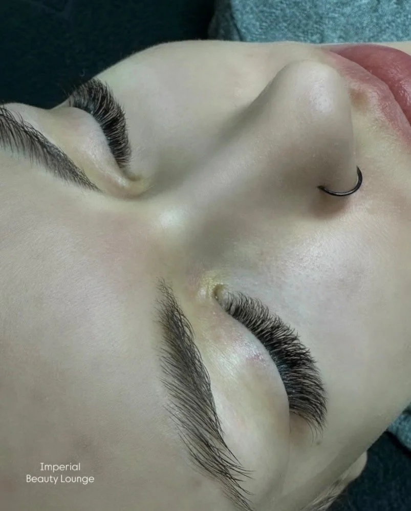 Close-up of a woman's face with freshly done eyelash extensions, a nose ring, and closed eyes, taken inside a beauty lounge.