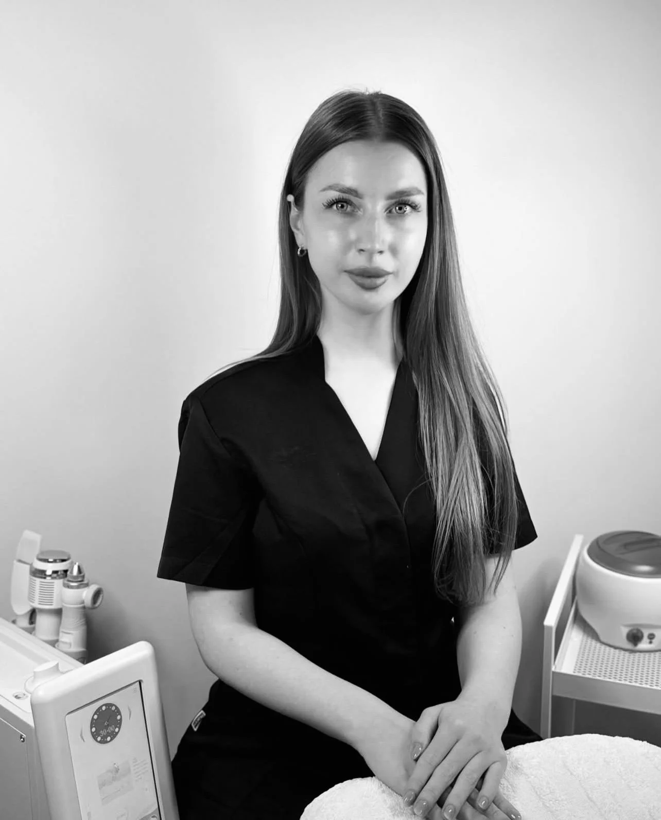 A woman with long hair and earrings wearing a black uniform, standing in a clinical or spa setting, with equipment and a towel nearby.