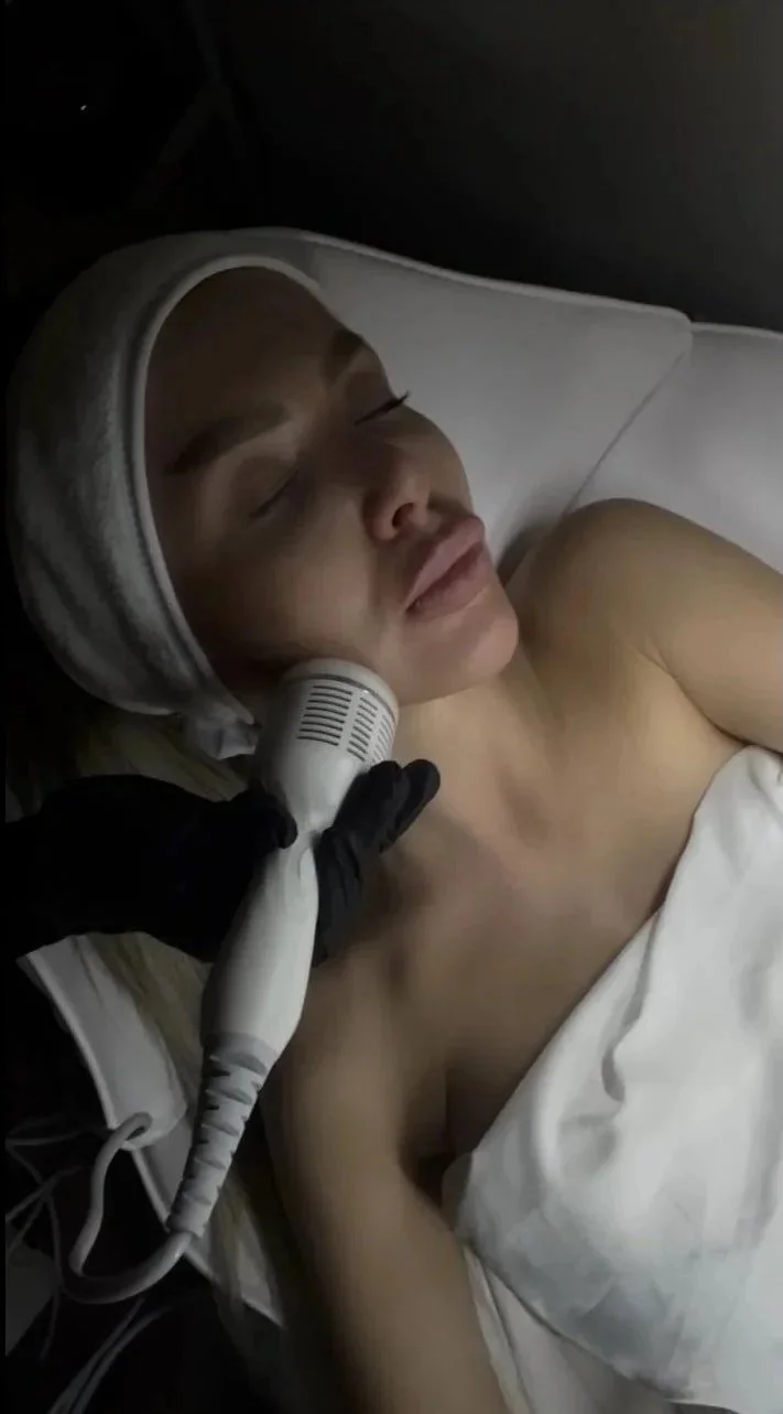 A woman with fair skin and a white headband is lying in a hospital bed with closed eyes. A gloved hand is holding a medical device against her cheek.