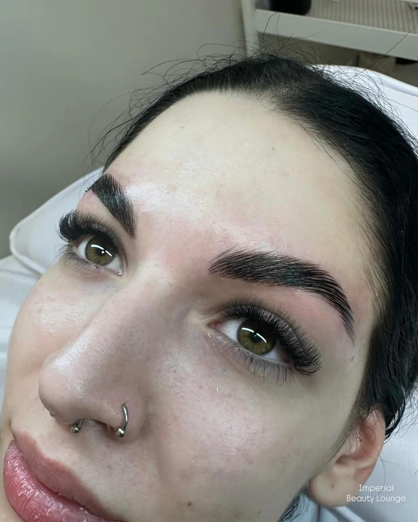 Close-up of a woman's face showing freshly shaped dark eyebrows, long eyelashes, and piercings on her nose, with a background indicating that she is at a beauty salon or clinic.