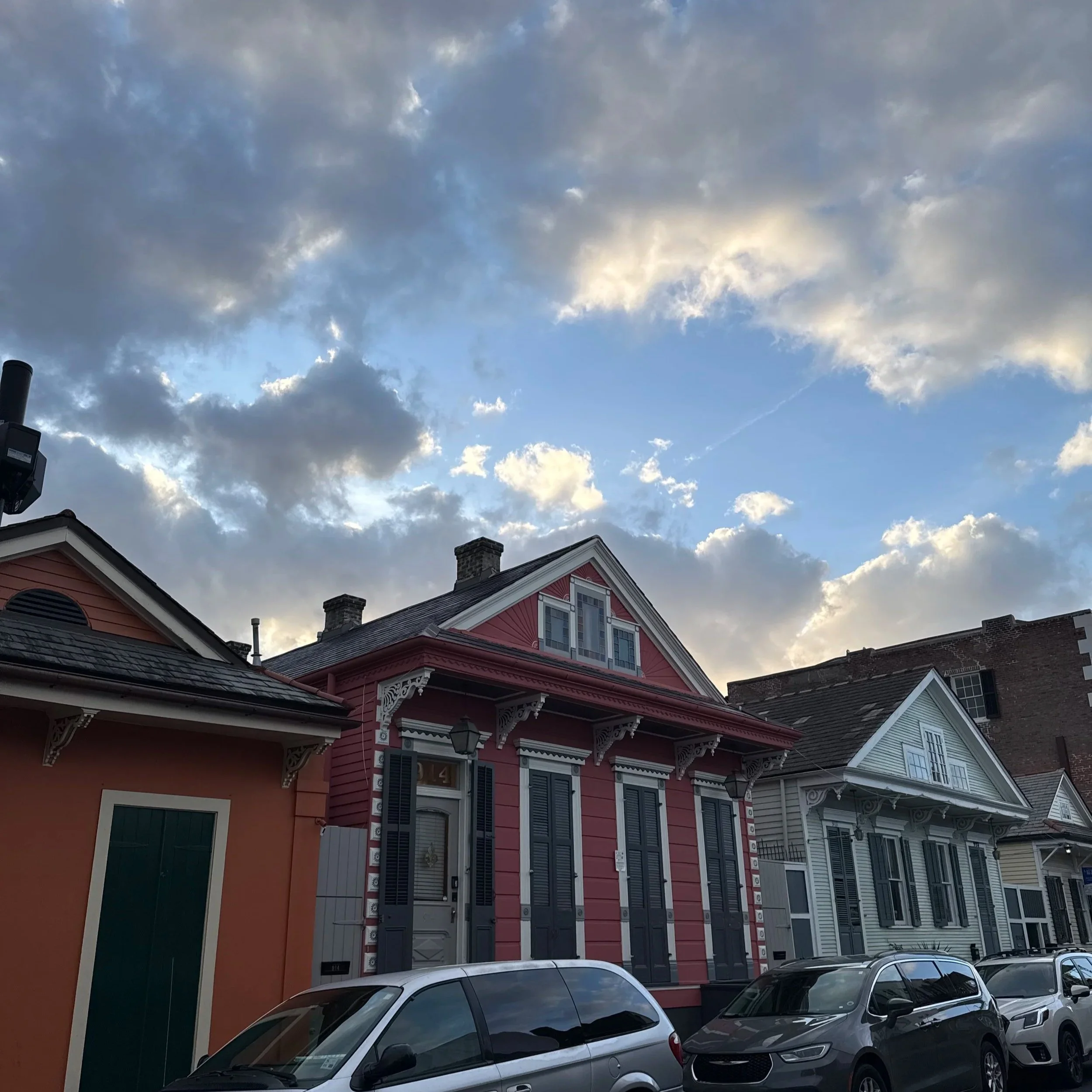 Colorful houses on a street with a cloudy sky overhead. The houses have Victorian-style architecture, with decorative trim and shutters, and are painted in pink, white, and orange. Cars are parked in front of the houses.