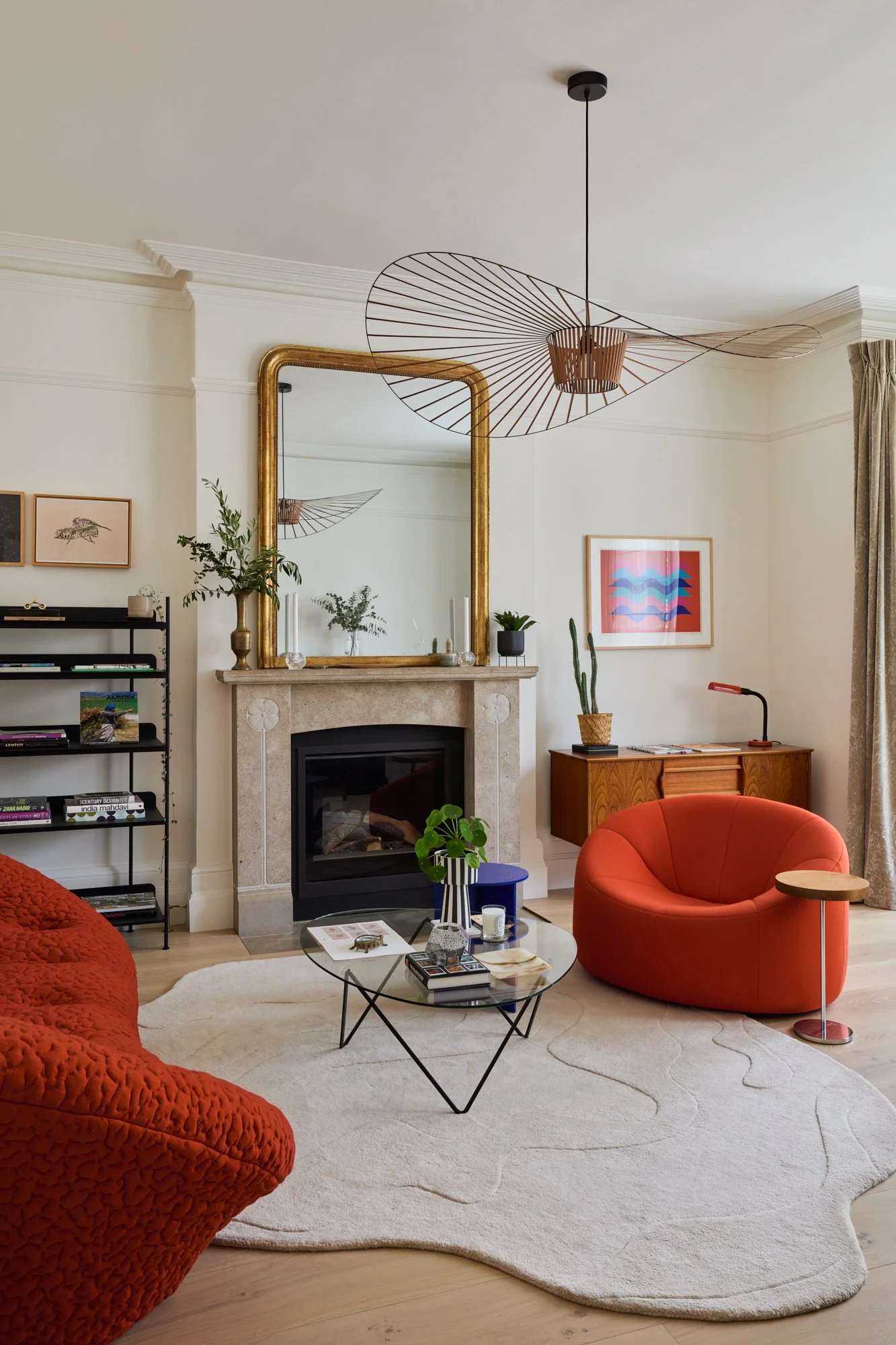Parisian-inspired formal reception room with high ceilings and large windows, styled with a Louis Philippe mirror, statement pendant, and French post-modern orange sofas. The design blends Haussmannian elegance with Scandinavian and mid-century moder