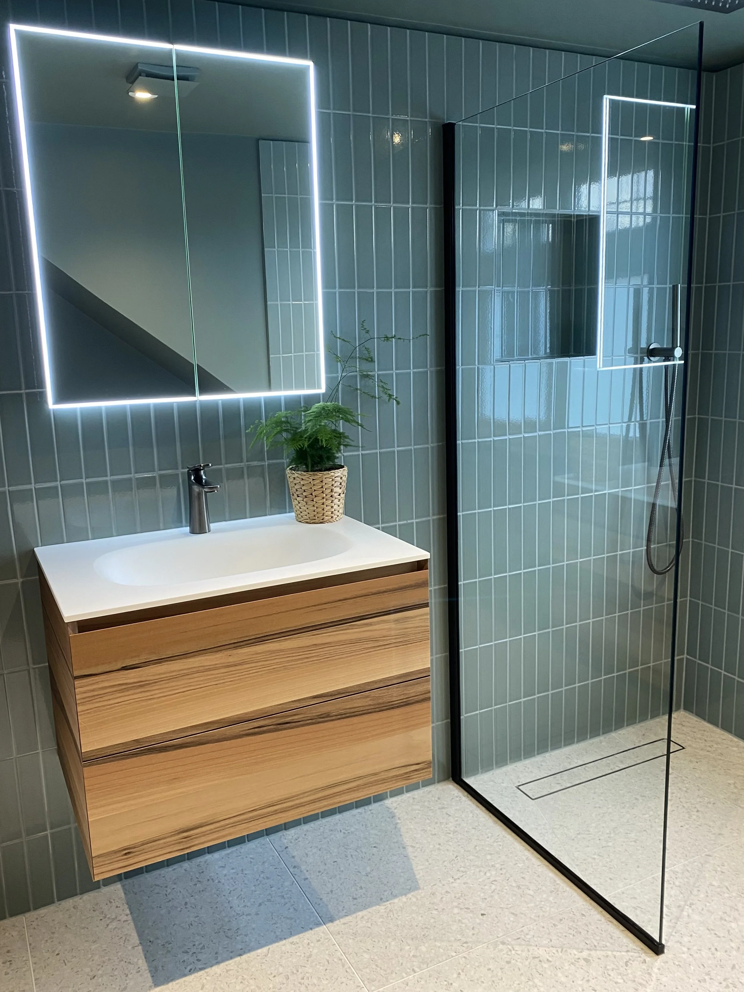 Compact bathroom addition under the eaves with colour-drenched warm blue/green walls, terrazzo flooring and vertical tiles, brushed black fixtures, and bespoke walnut vanity with Corian top.