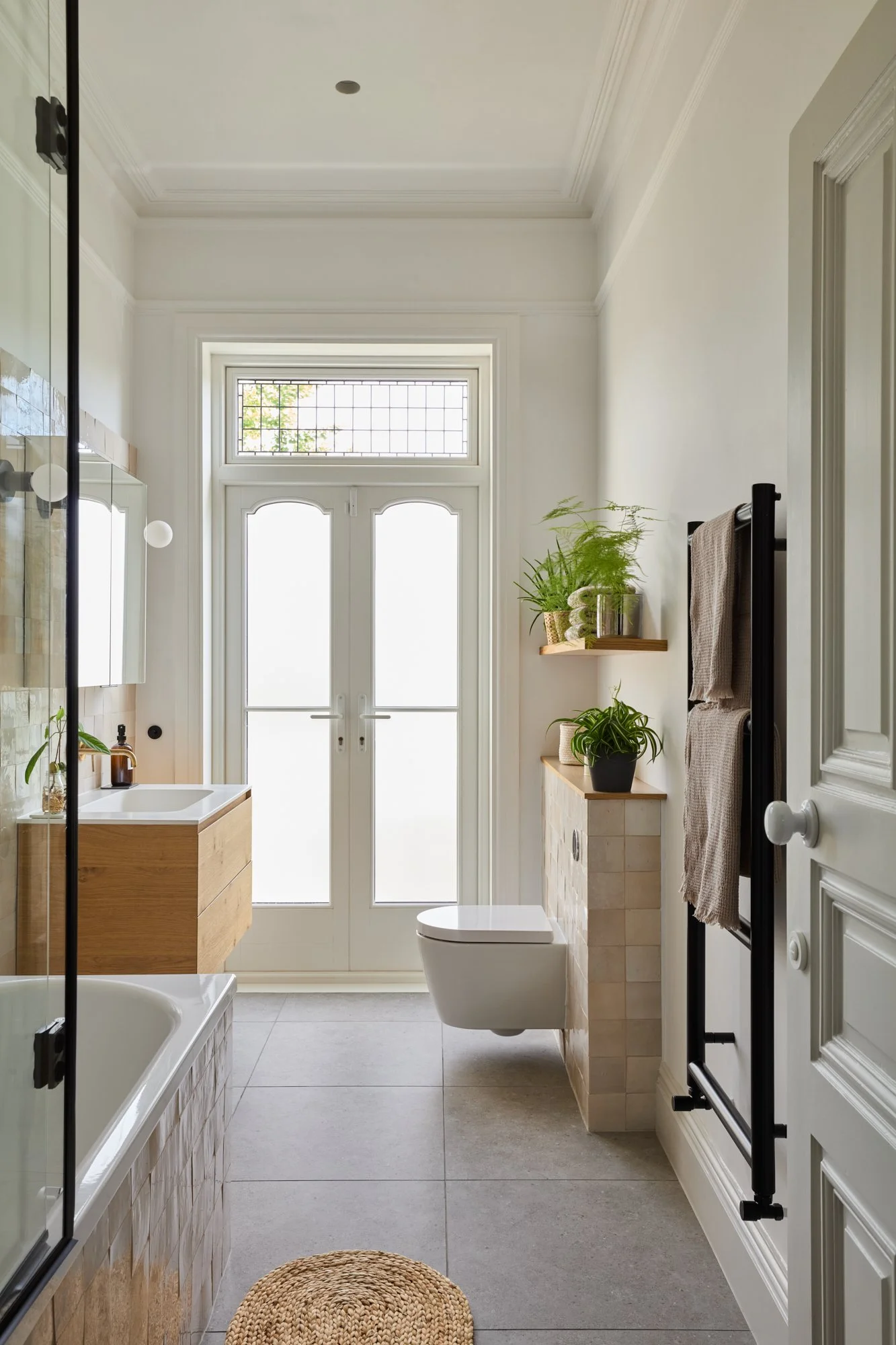 This calm family bathroom combines handmade zellige tiles, aged brass fixtures, and light oak joinery. Double glass doors bring natural light, while layered greenery adds a fresh touch to the warm neutral palette.