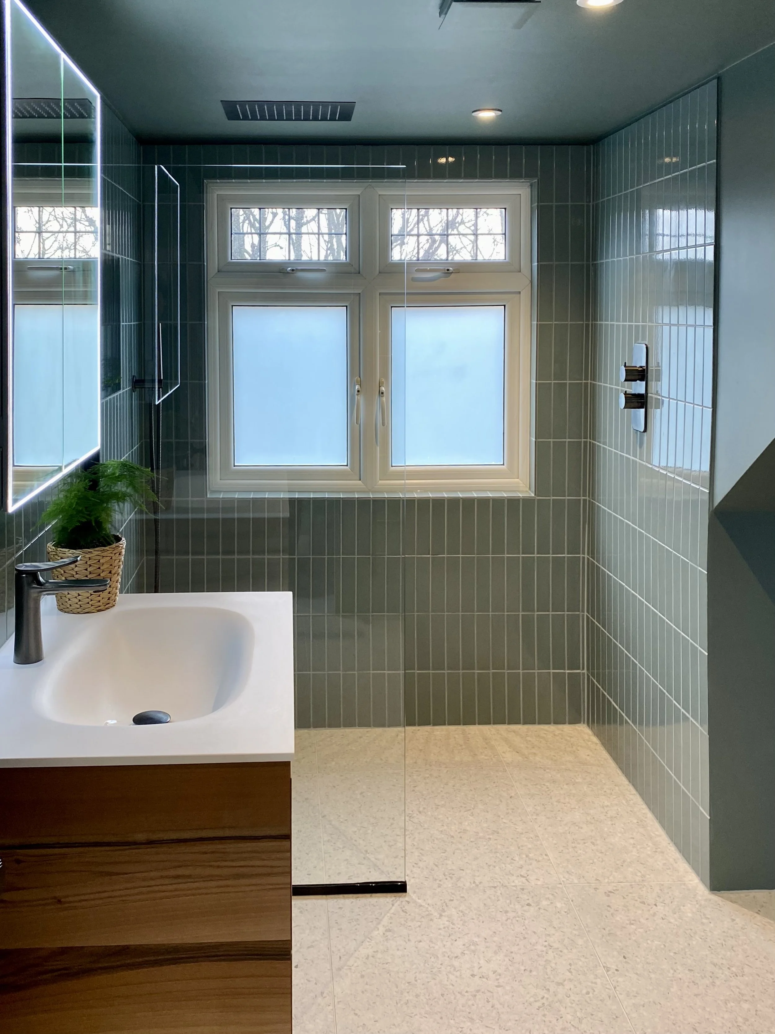 Small bathroom under the eaves with colour-drenched warm blue/green walls, terrazzo flooring and vertical tiles, brushed black fixtures, and bespoke walnut vanity with Corian top.