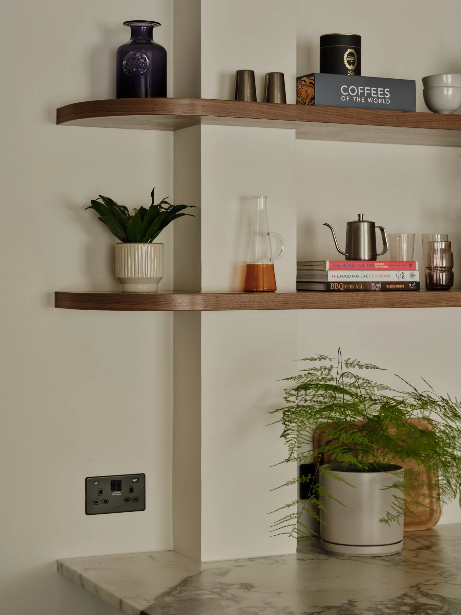 Open walnut kitchen shelves styled with plants, cookbooks and ceramics above Arabescato marble worktops in a Fulham kitchen.