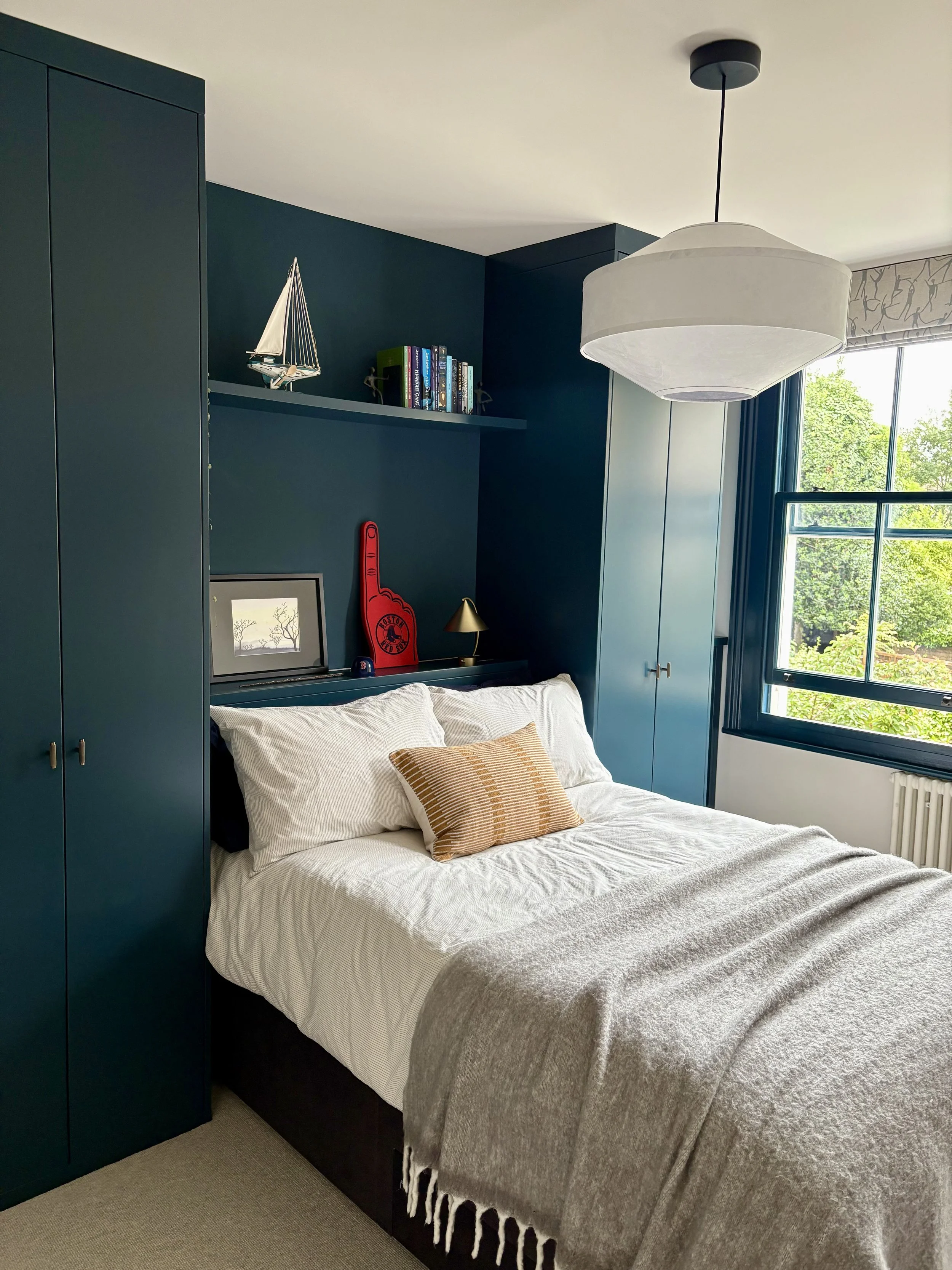 Modern teen bedroom in Clapham featuring bespoke dark blue fitted wardrobes, a recessed headboard with integrated shelving, and a large statement pendant.