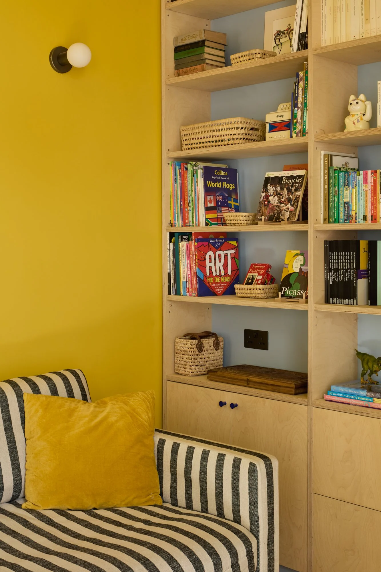 Vibrant playroom and family room with yellow, red, and blue accents. Features a chimney breast desk nook, bespoke birch plywood joinery, PH5 pendant, stripy sofa, jute rug, and decorative plants for a playful yet functional space.