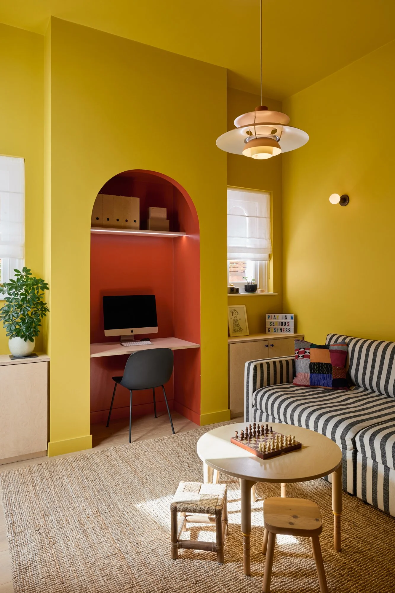 Vibrant playroom and family room with yellow, red, and blue accents. Features a chimney breast desk nook, bespoke birch plywood joinery, PH5 pendant, stripy sofa, jute rug, and decorative plants for a playful yet functional space.