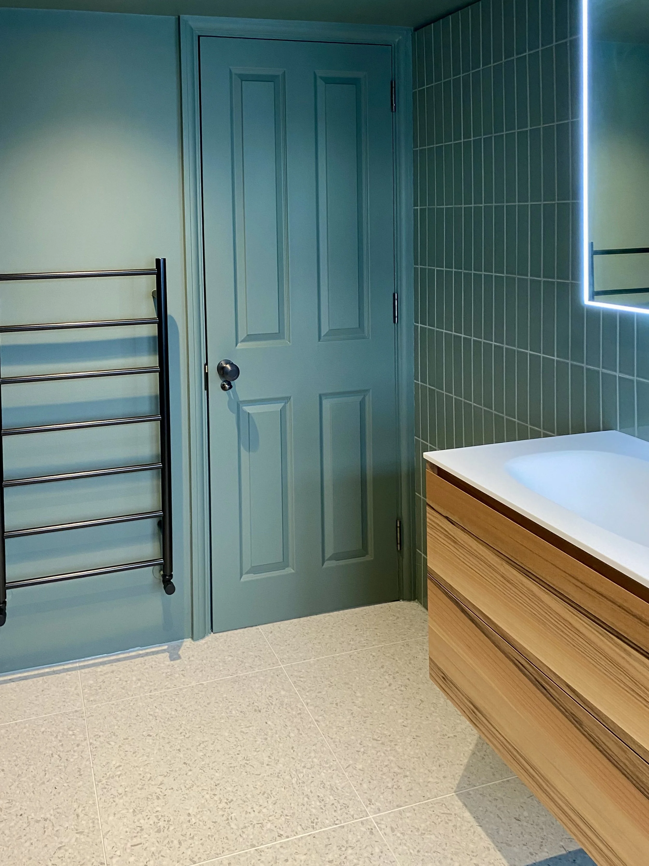 Compact bathroom addition under the eaves with colour-drenched warm blue/green walls, terrazzo flooring and vertical tiles, brushed black fixtures, and bespoke walnut vanity with Corian top.