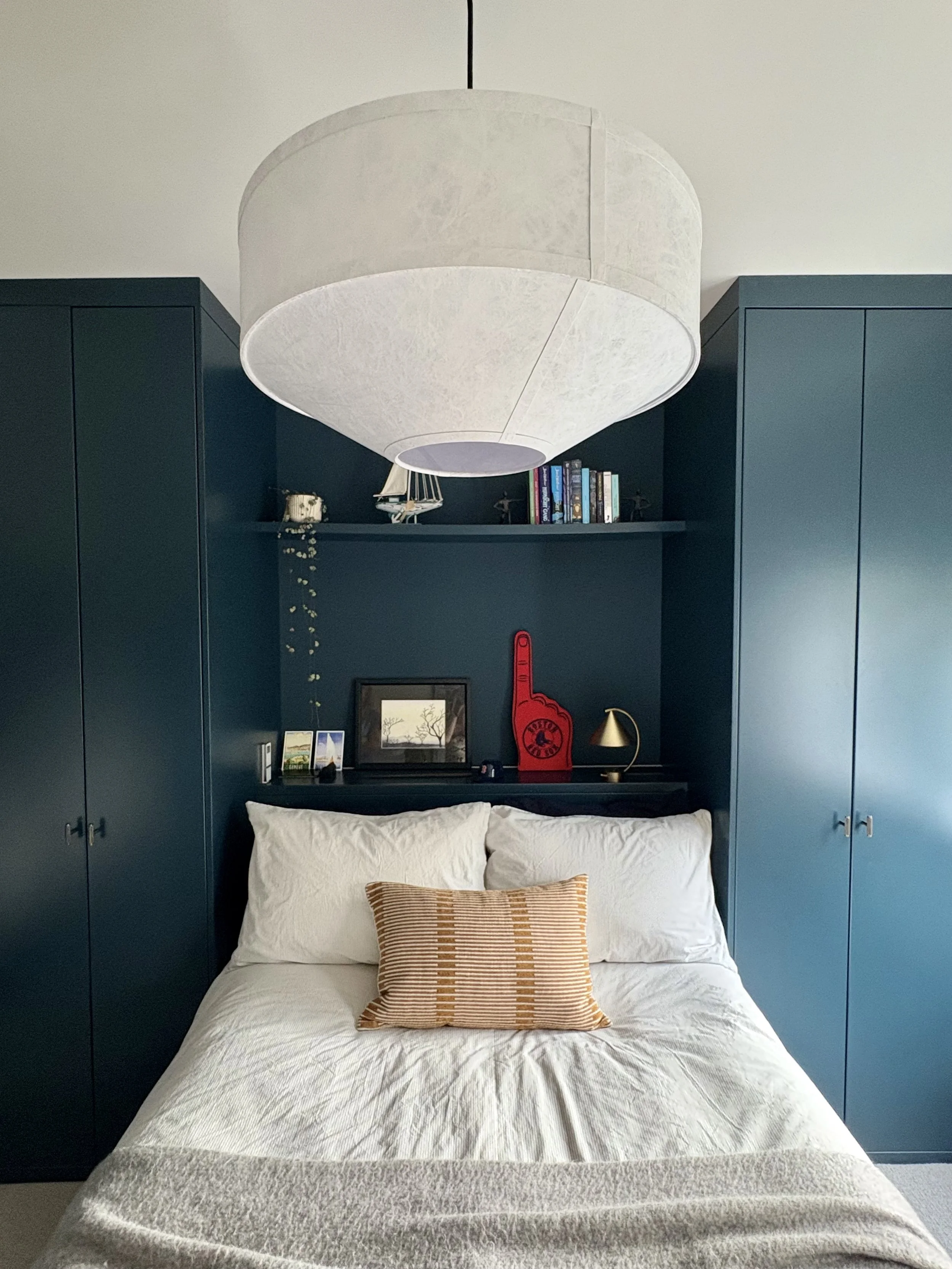 Modern teen bedroom in Clapham featuring bespoke dark blue fitted wardrobes, a recessed headboard with integrated shelving, and a large statement pendant.